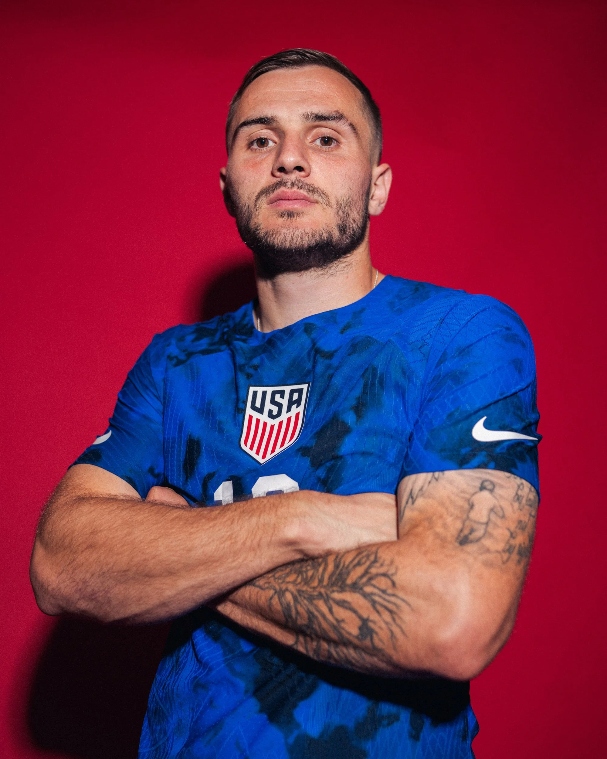 A man with a beard and tattoos on his arms standing with arms crossed wearing a blue USA soccer jersey against a red background.