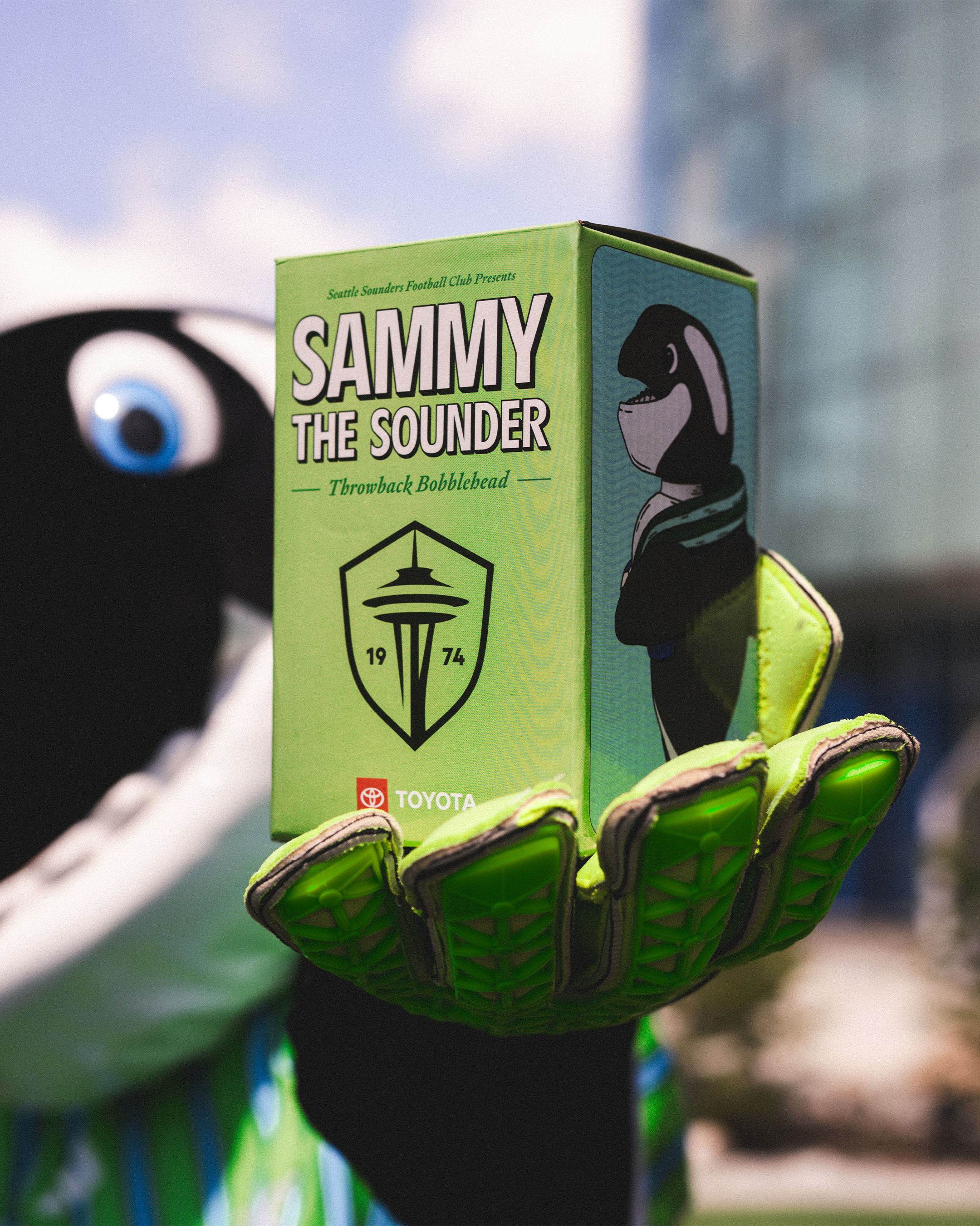 Close-up of a green box labeled 'Sammy the Sounder' held in a mascot's gloved hand. The mascot has an animated black and white penguin wearing a hoodie. The box features a logo of the Seattle Seahawks and Toyota logo at the bottom.