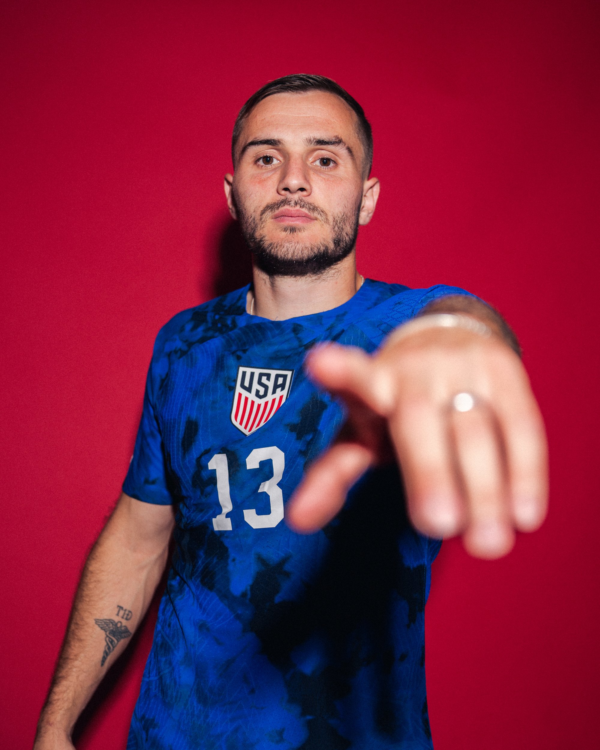 A man in a blue USA soccer jersey with the number 13 pointing towards the camera, standing against a red background.