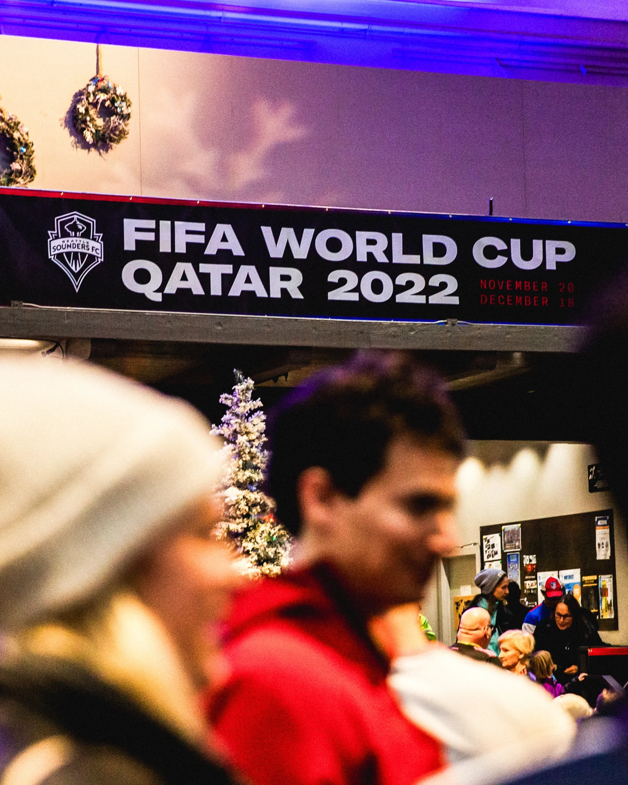 A crowded indoor venue with a sign displaying FIFA World Cup Qatar 2022, dates November 20 to December 18, 2022. There are people in the foreground, some wearing winter clothing, and a decorated Christmas tree is visible in the background.
