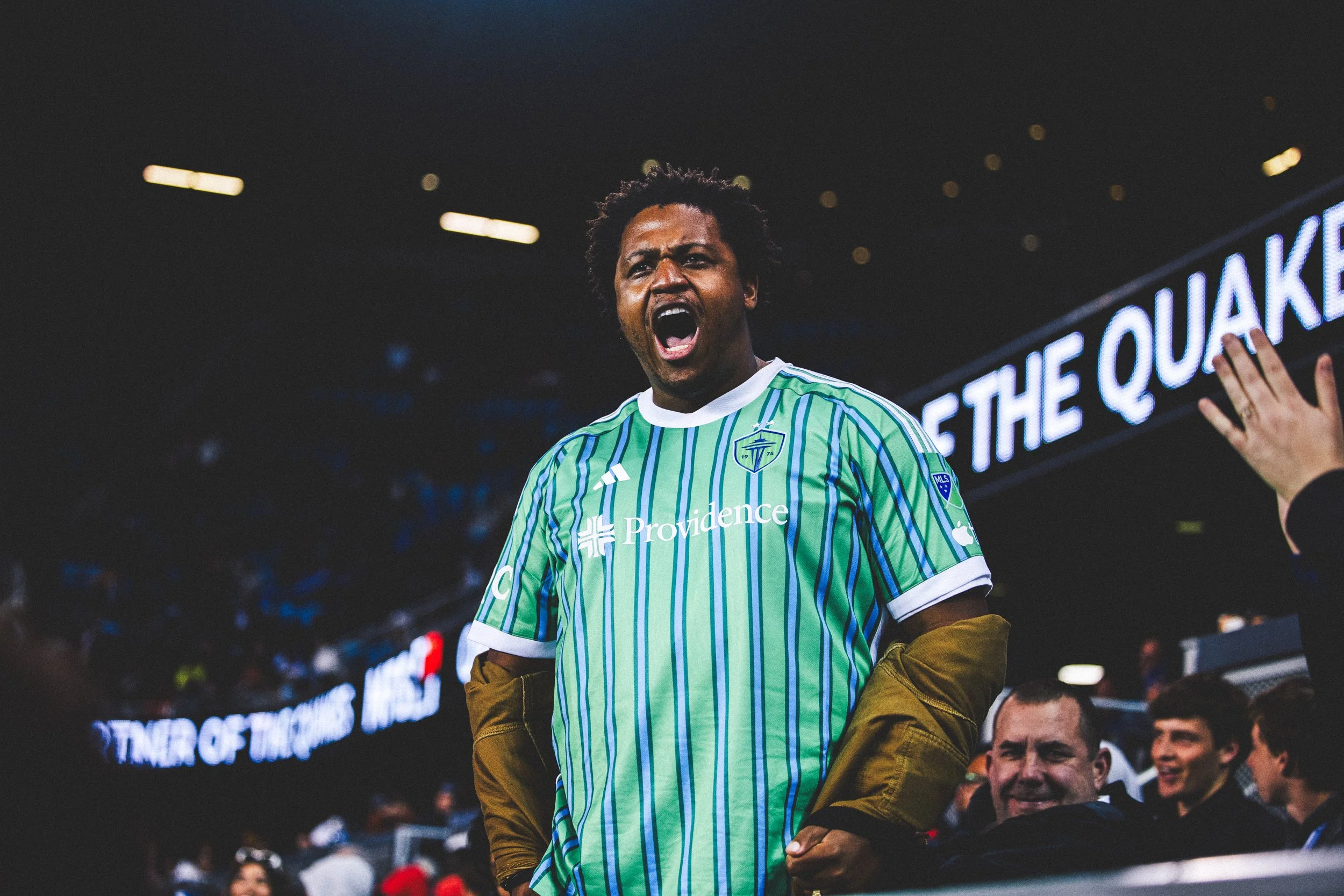 A man in a green and blue striped soccer jersey yelling passionately at a sports event.