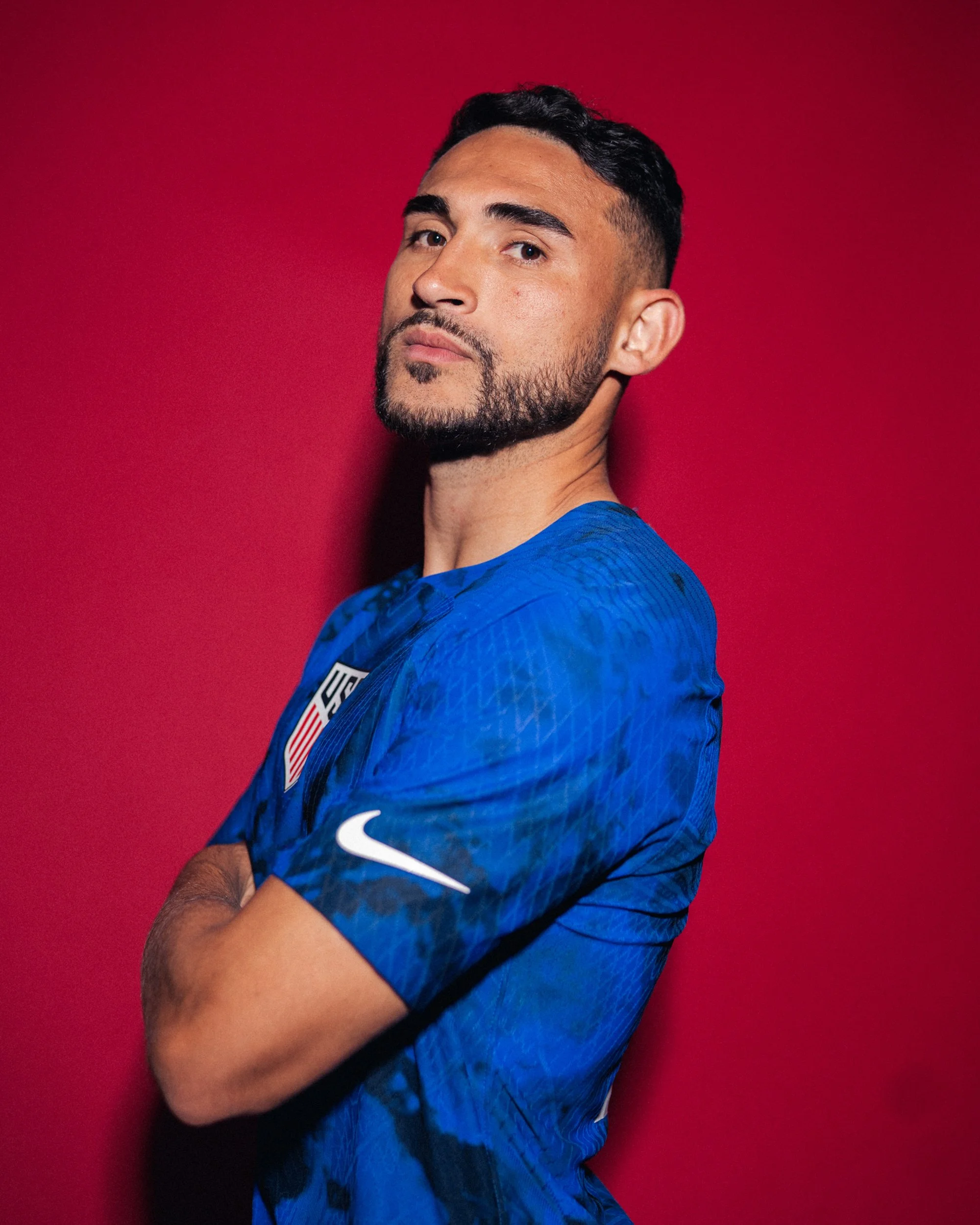 A man in a blue sports jersey standing with arms crossed against a red background.