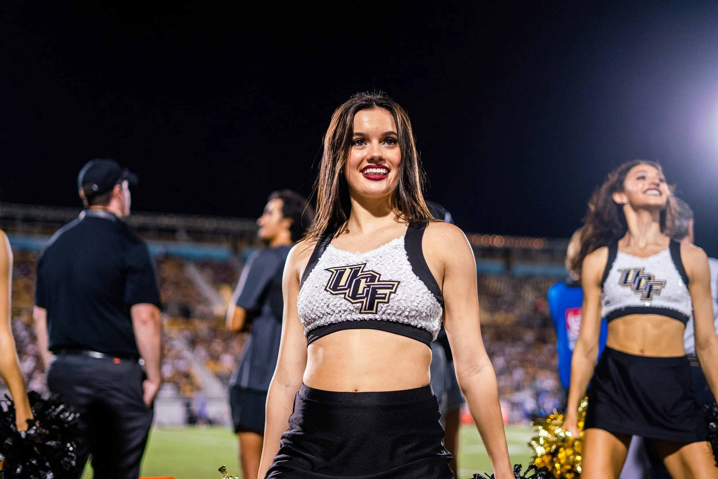 Cheerleaders performing at a nighttime sports event, wearing UCF uniforms and black skirts, with a stadium and crowd in the background.