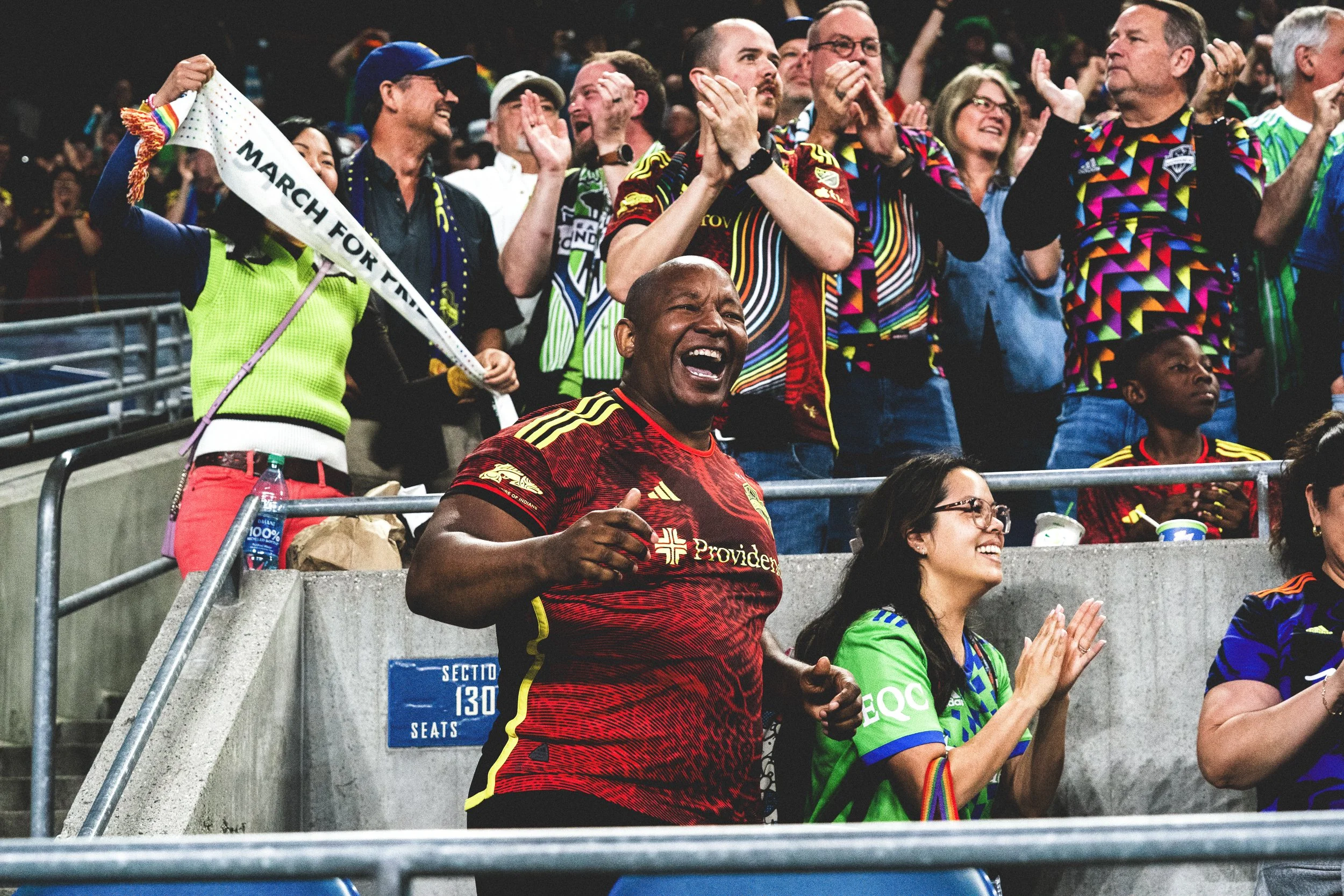 Crowd of soccer fans celebrating, some wearing colorful jerseys and holding scarves, with one person holding a "March for Unity" banner, at a stadium.