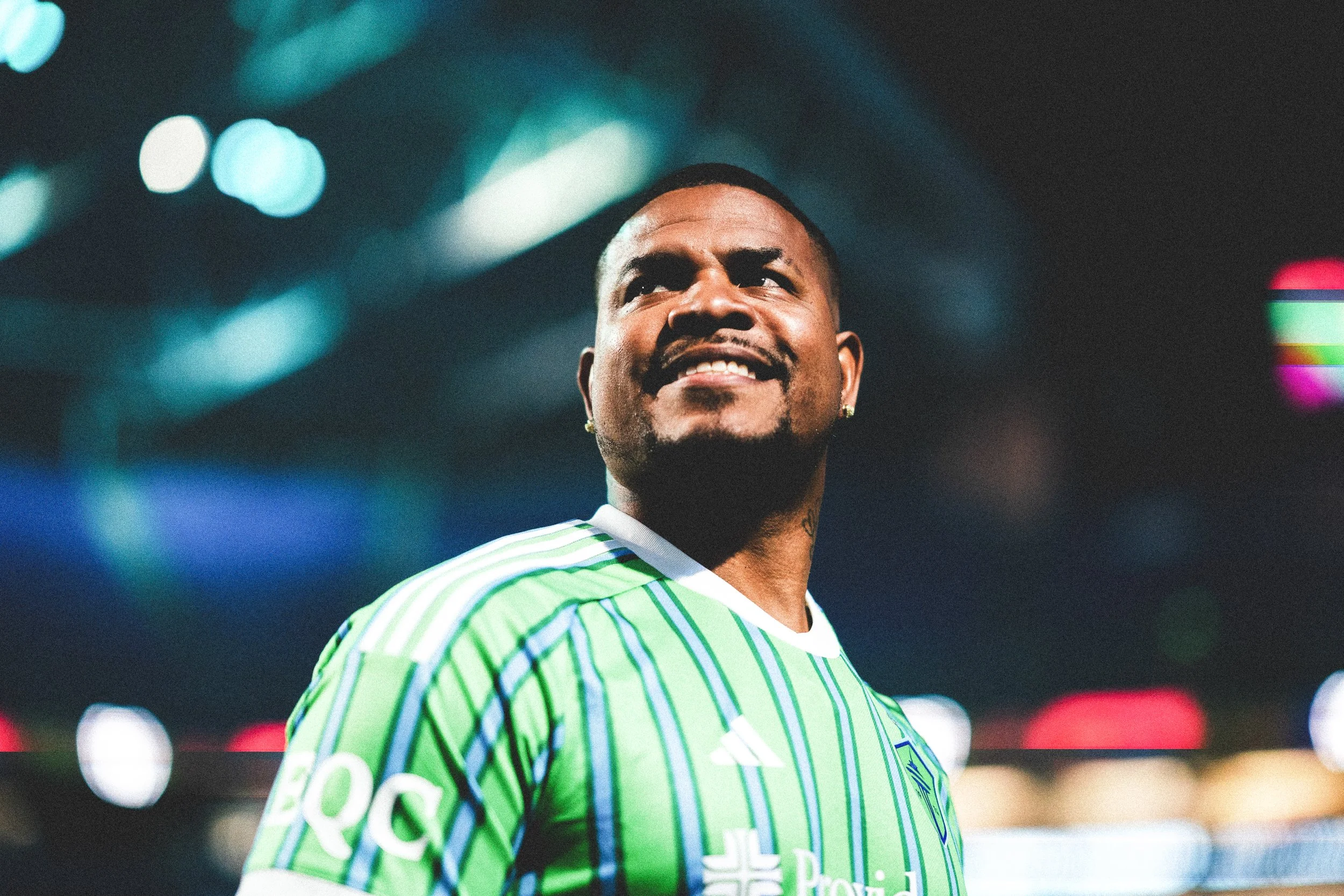 A male soccer player in a green and white striped jersey, smiling and looking upward, with a blurred stadium background illuminated by colorful lights at night.
