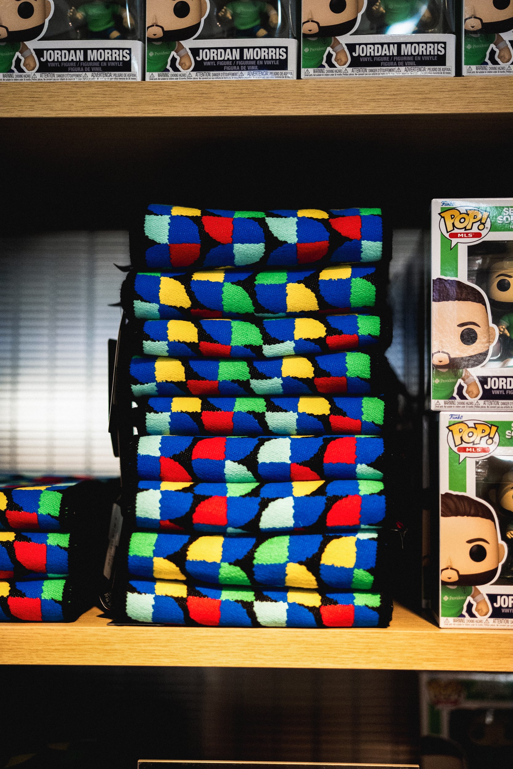 Stack of colorful socks with geometric patterns arranged on a wooden shelf, with Jordan Morris Funko Pop figures on the right and box of Jordan Morris Funko figures on the top shelf.