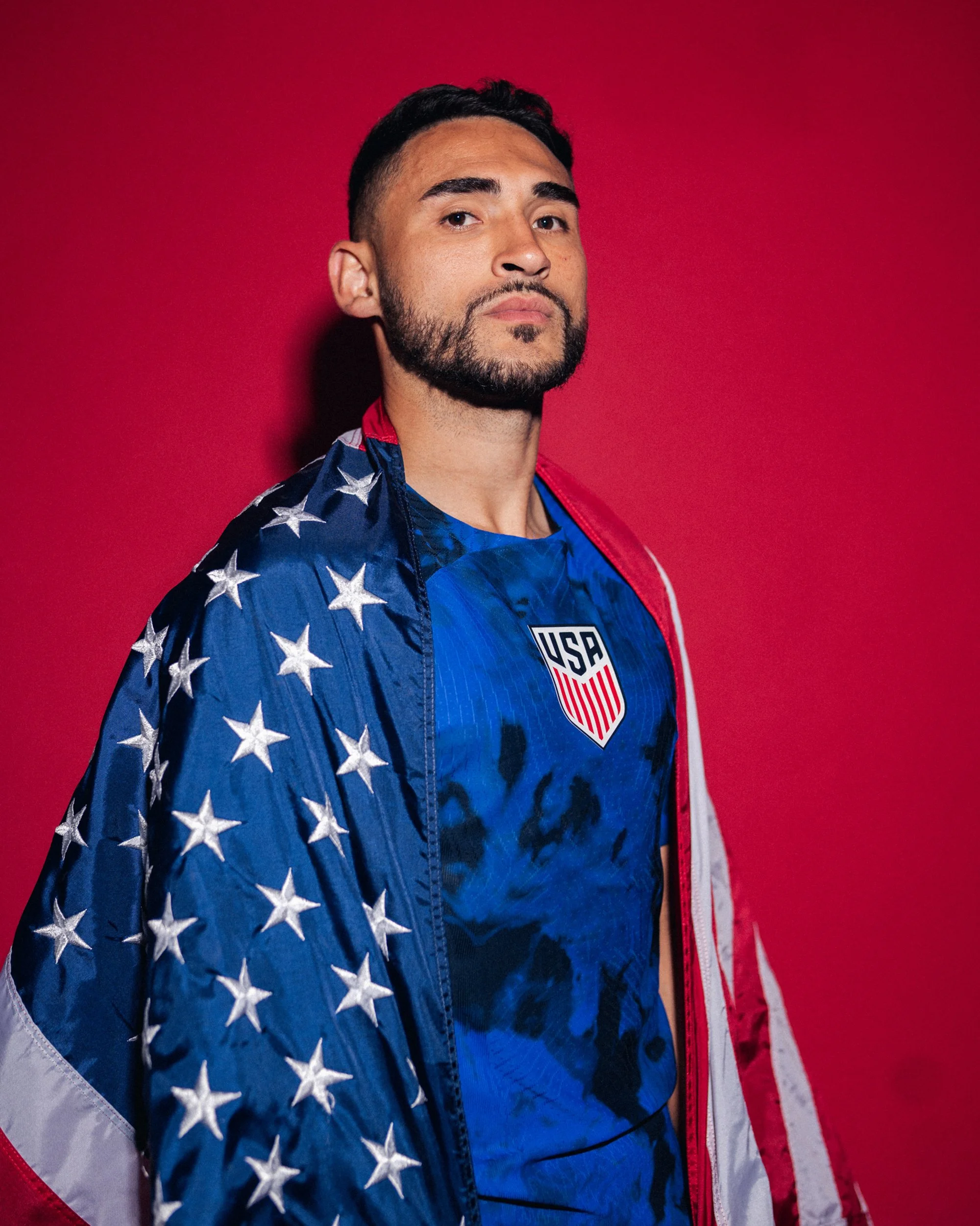Man wearing USA athletic jersey and jacket with American flag design, standing against red background.