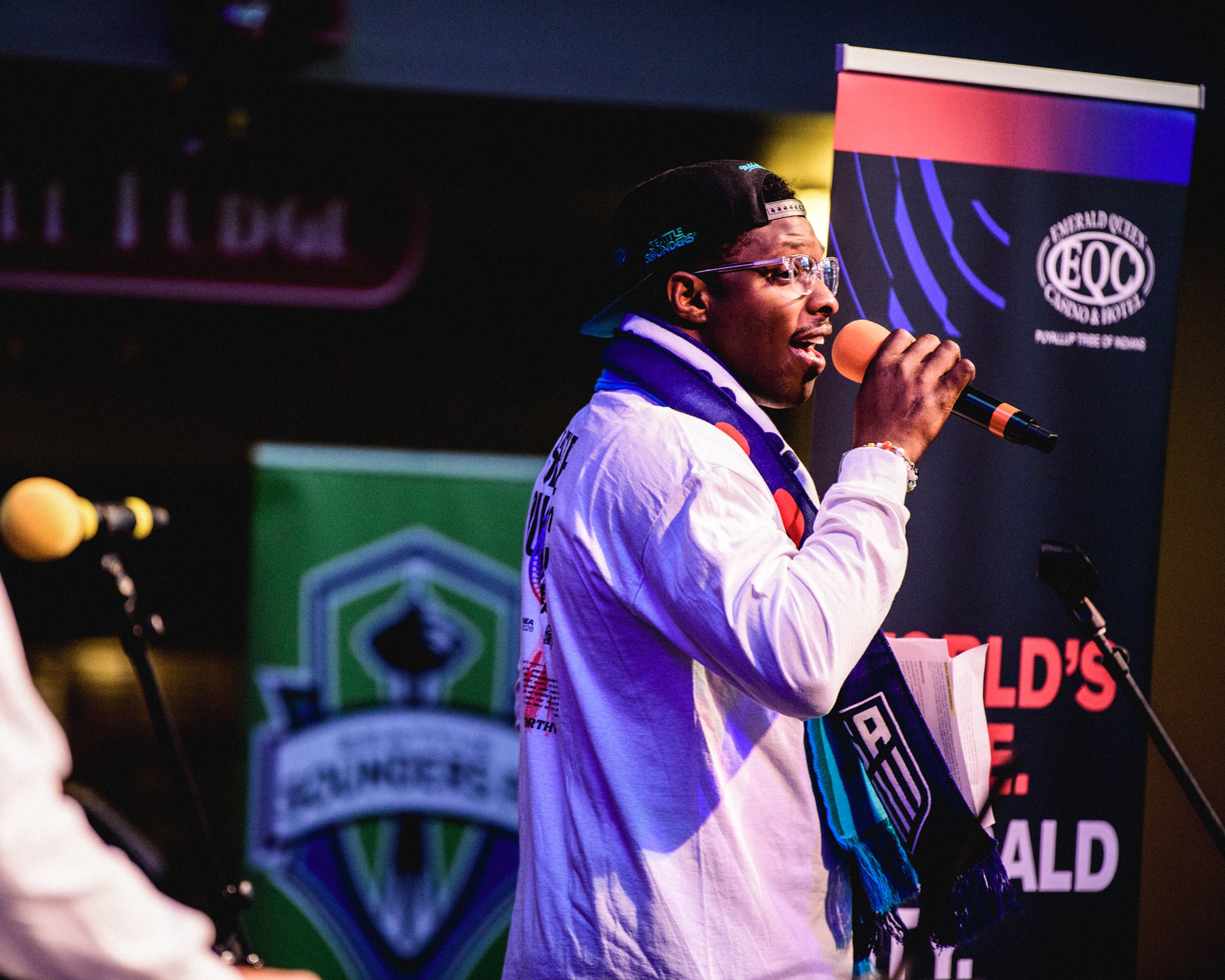 A man wearing a black cap backwards, clear glasses, a white jacket, and a scarf, speaking or singing into a microphone at an event with banners and microphones in the background.