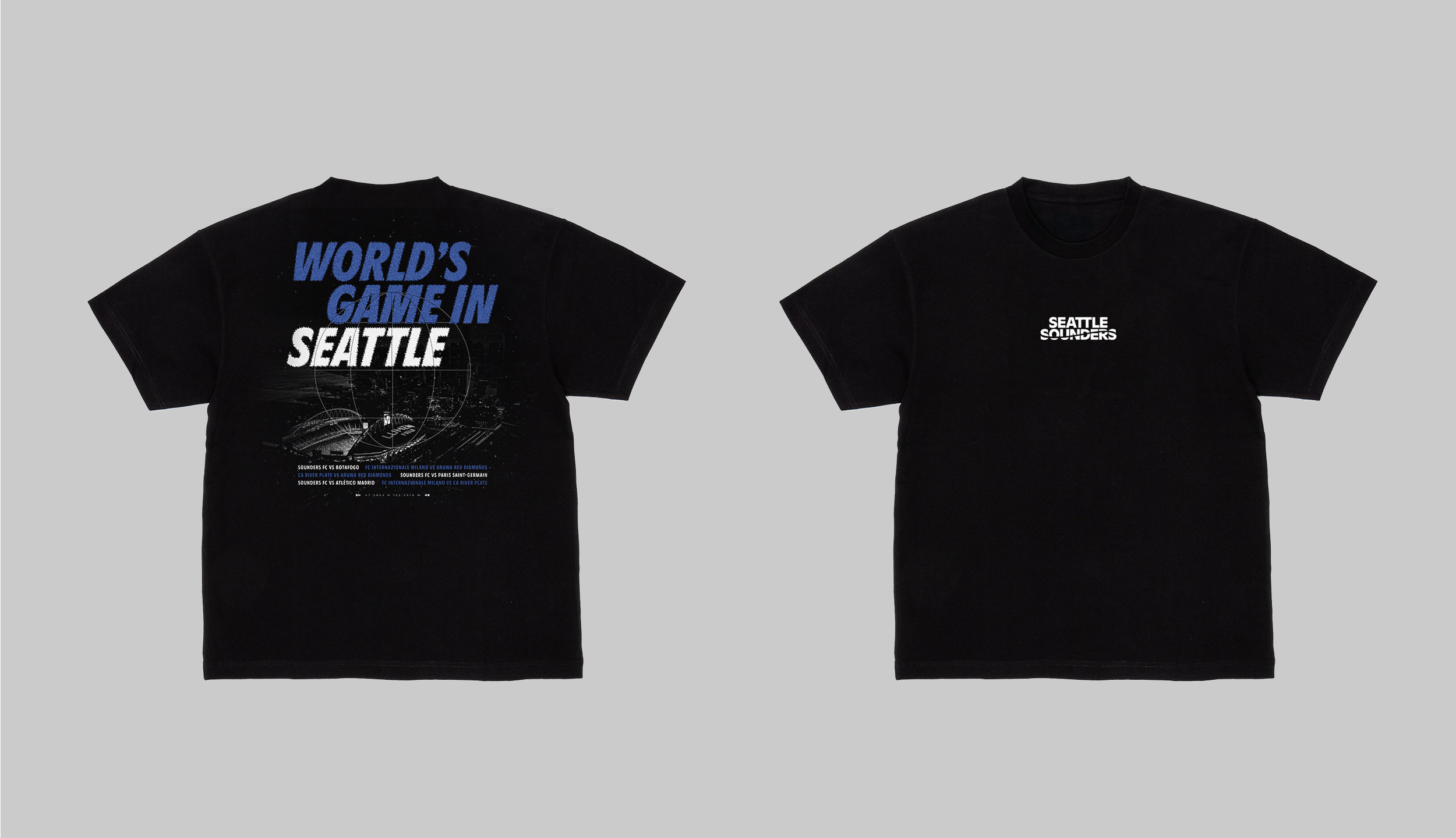Two black t-shirts, one with a large graphic design and the other with small text. The left shirt features the text "WORLD'S GAME IN SEATTLE" in blue and white, with a detailed illustration of a stadium and cityscape underneath. The right shirt has t