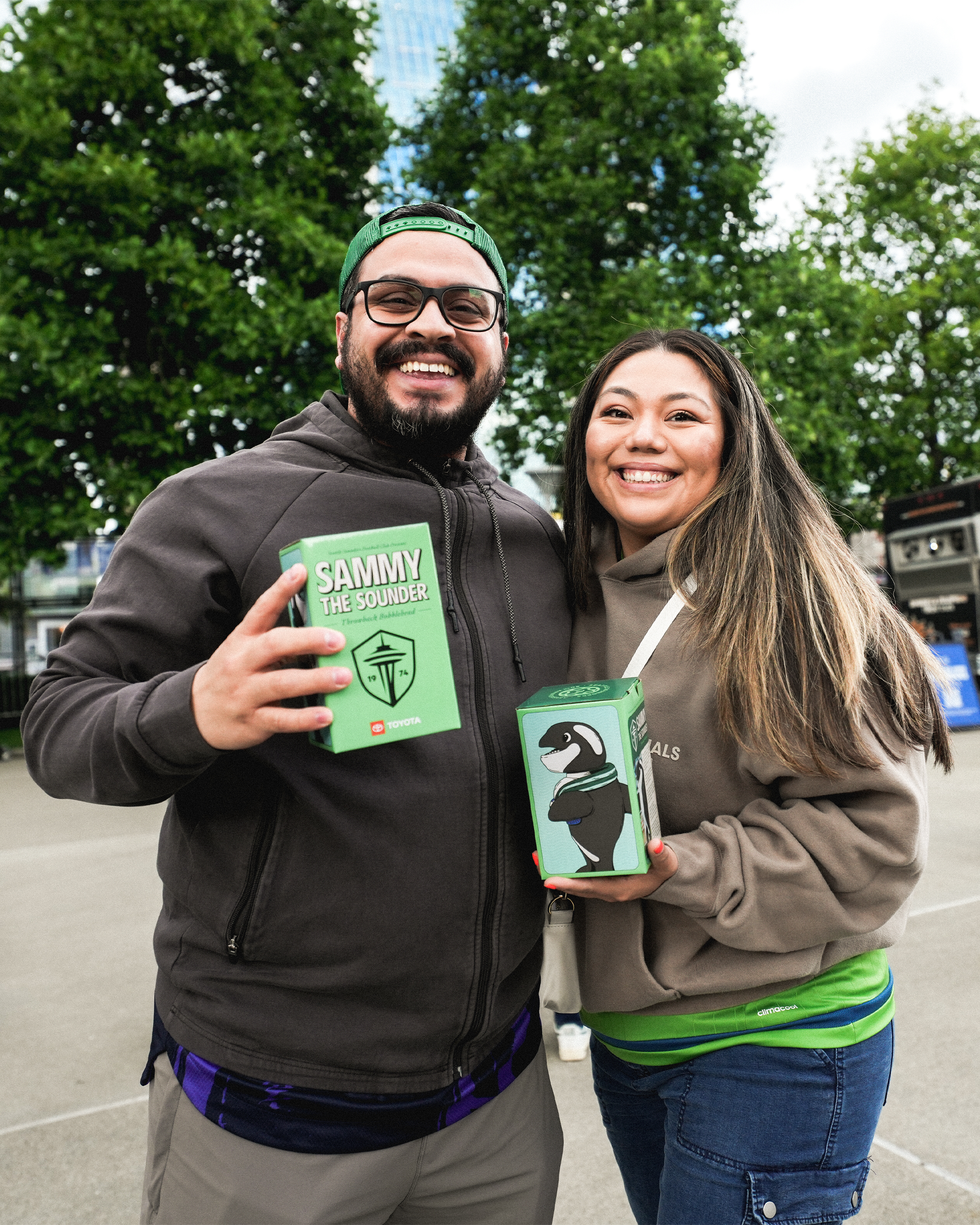 Two smiling people holding square green boxes in an outdoor setting with trees and a bus background.