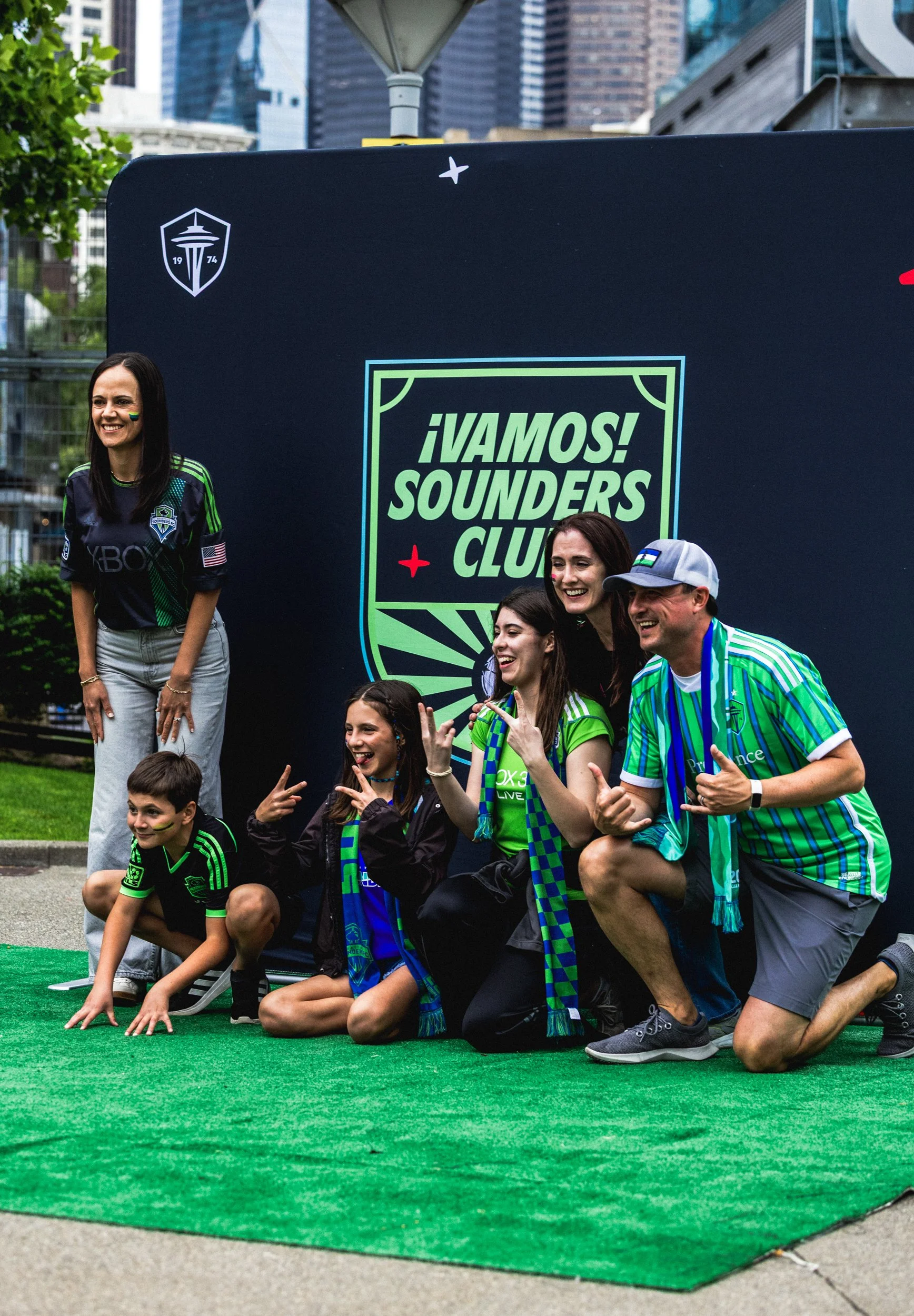 Group of people posing in front of a large sign that reads '¡Vamos! Sounders Club' at an outdoor event, with some wearing Seattle Sounders soccer team apparel.