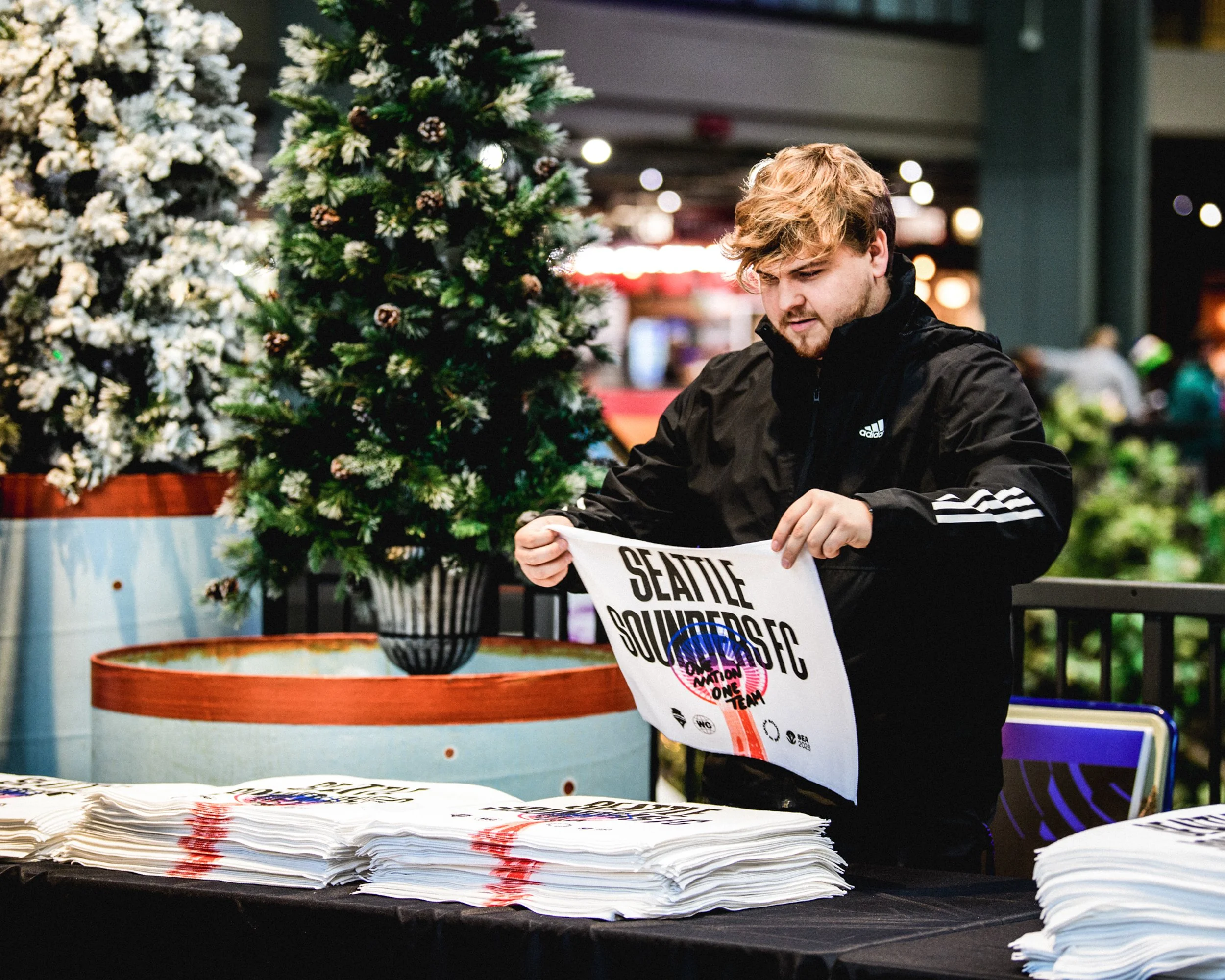 A young man in a black Adidas jacket organizing on a table that has stacks of white tote bags printed with an event logo, with Christmas trees and colorful lights in the background.