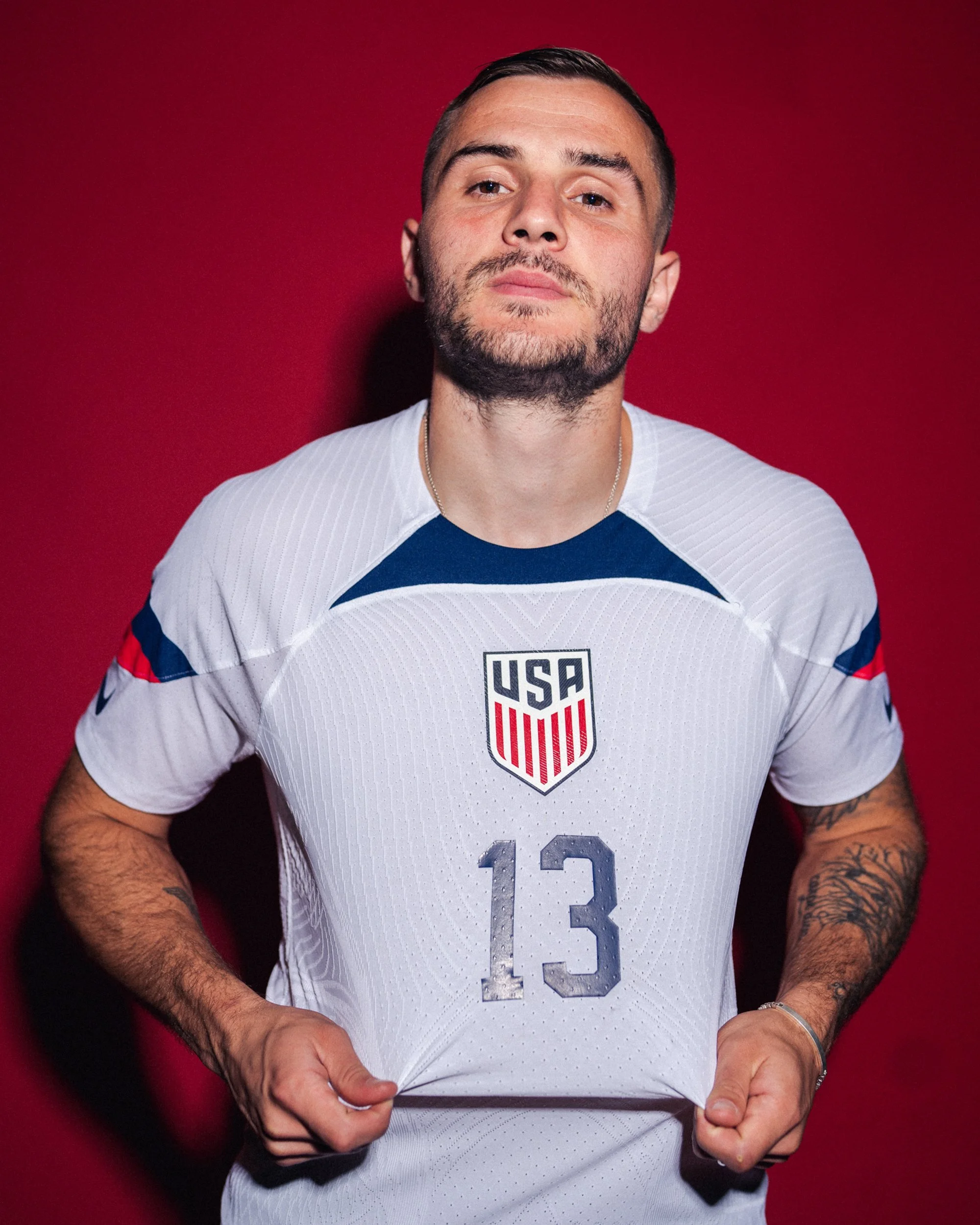 A man wearing a white USA soccer jersey with the number 13 and a USA emblem, standing against a red background.