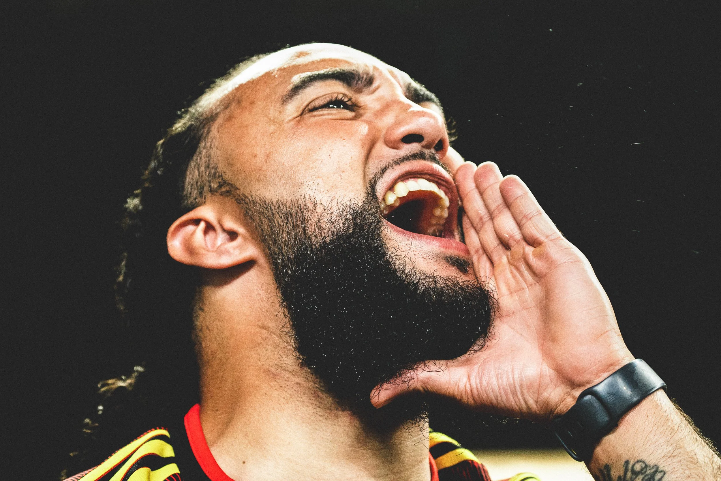 A man shouting with his hand near his mouth, wearing a sports uniform and a fitness tracker on his wrist.