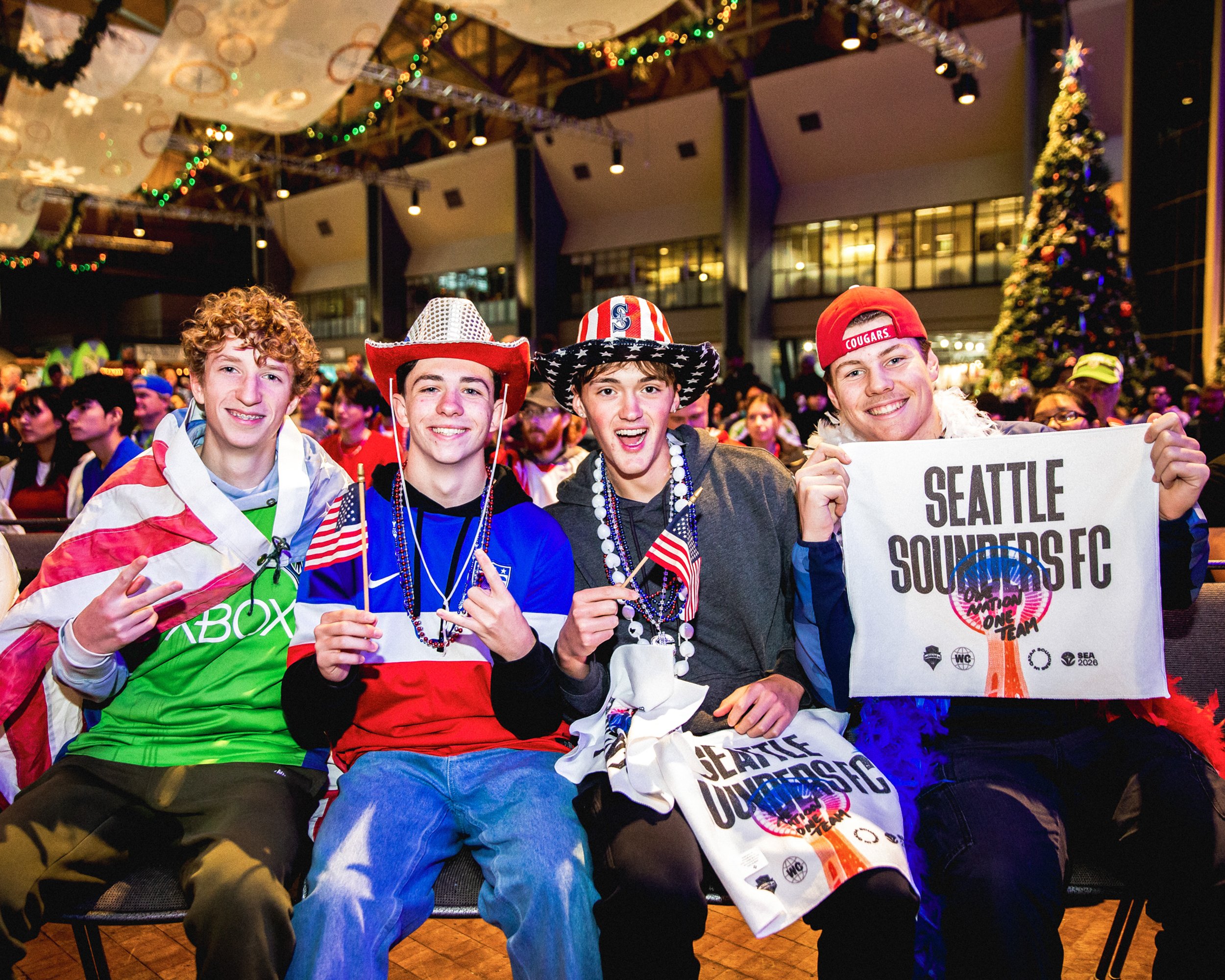 Four young men sitting together at an indoor event, dressed in patriotic and sports fan attire, holding small American flags and a Seattle Sounders FC banner, with a large decorated Christmas tree in the background and colorful lights hanging from th