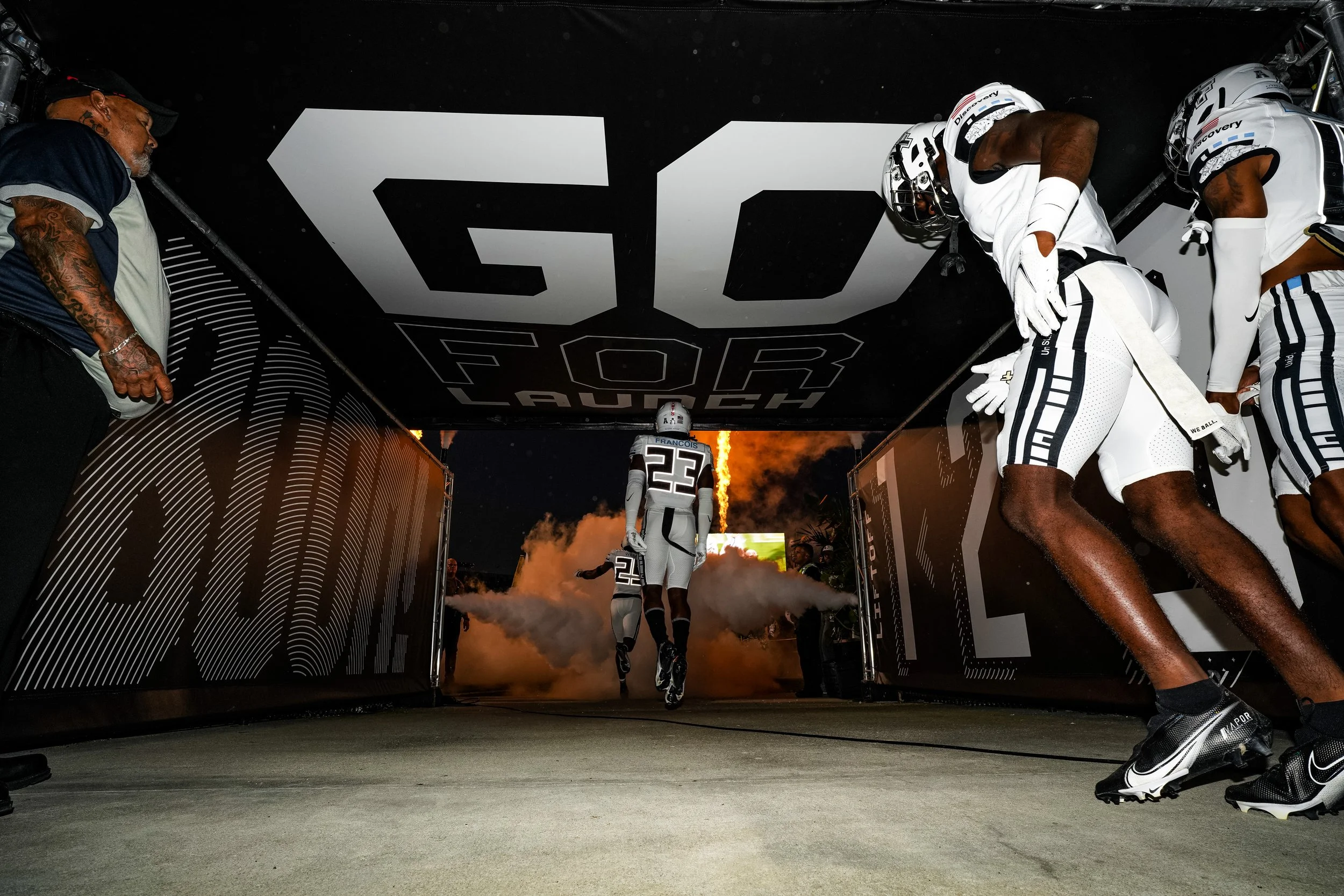 Football players running onto field through tunnel with smoke, with Fireworks in the background, under a sign that reads 'GOD FORFAUCH'.