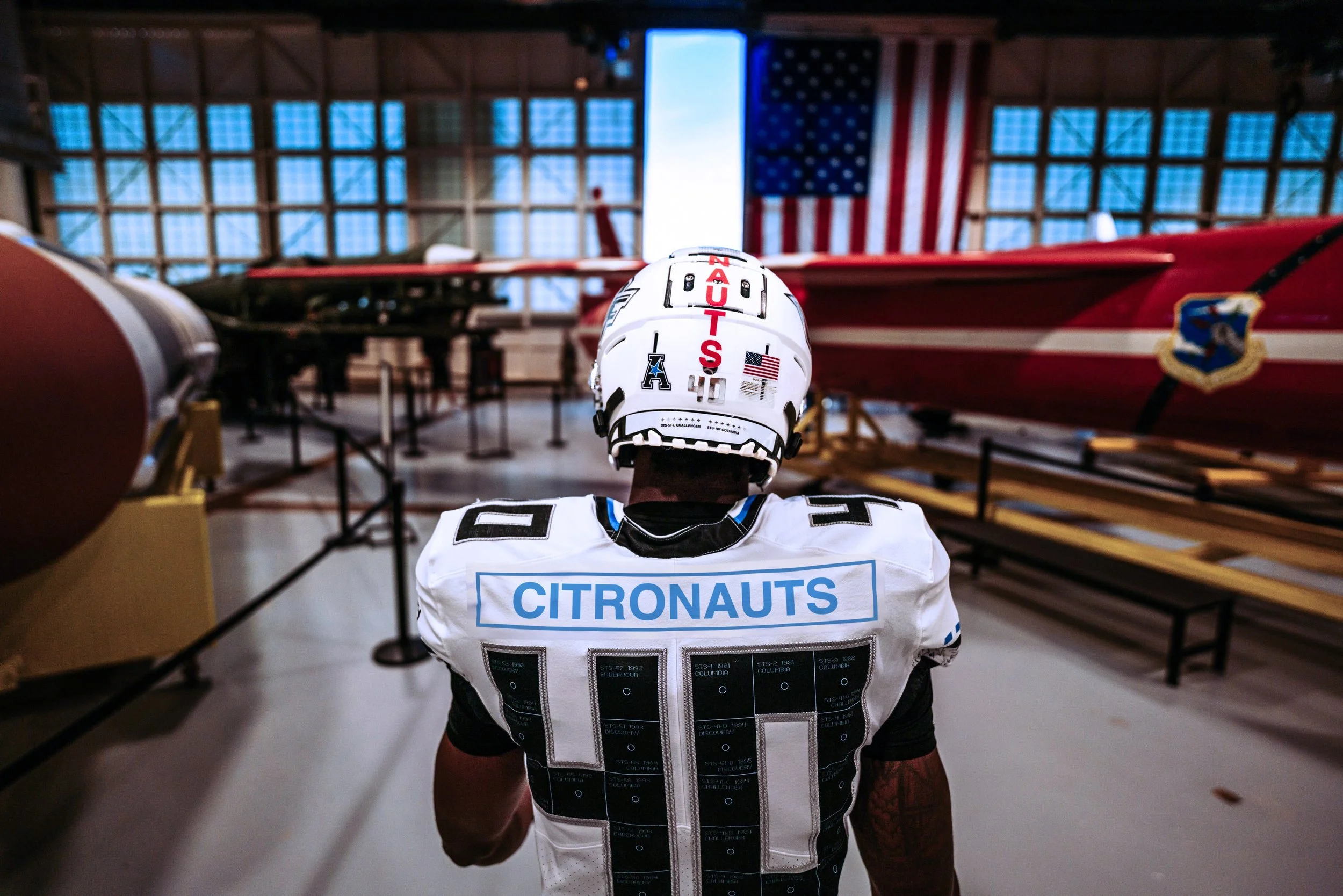 A football player in a white jersey with the word 'CITRONAUTS' on the back, wearing a helmet with the name 'NAUTS' and an American flag sticker, standing inside a museum with vintage aircraft and an American flag in the background.