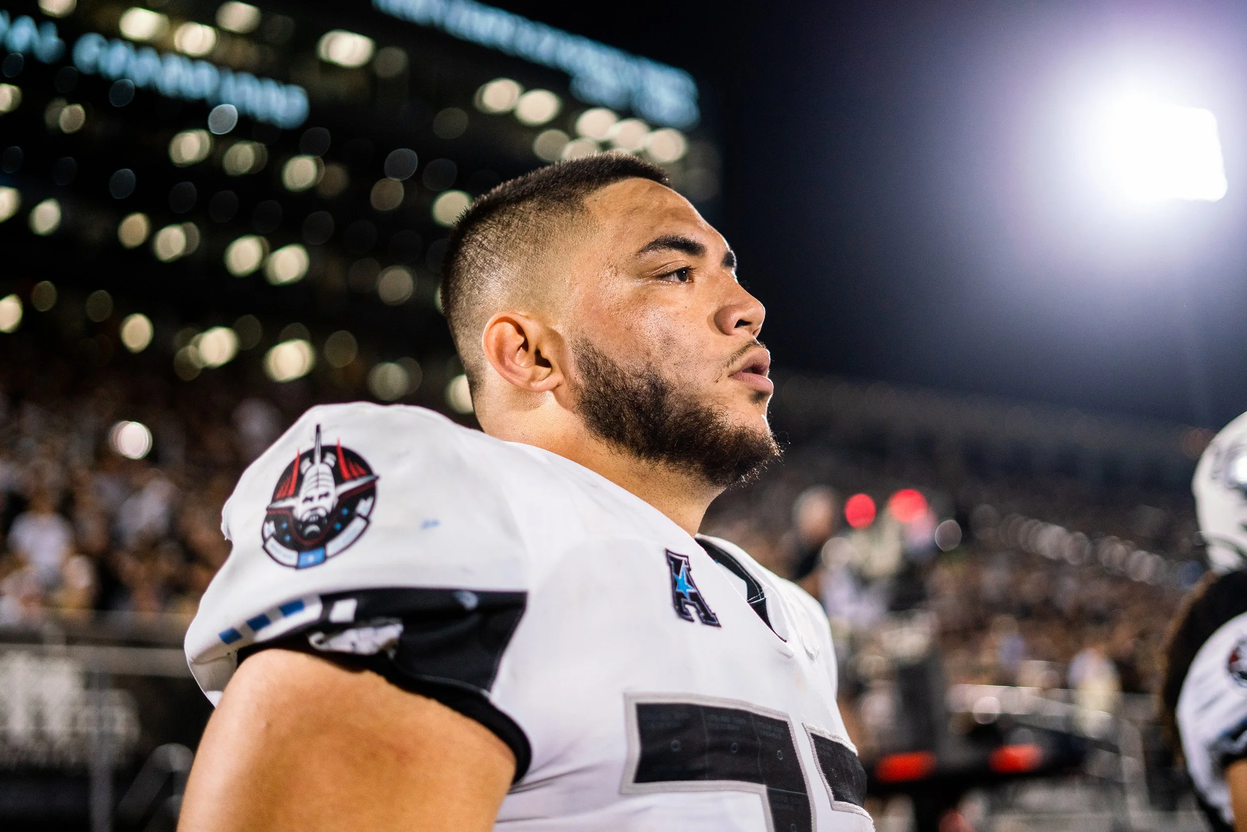 A professional football player with a beard and short hair, wearing a white jersey with black and blue accents, standing on the field during a night game with stadium lights and blurred spectators in the background.