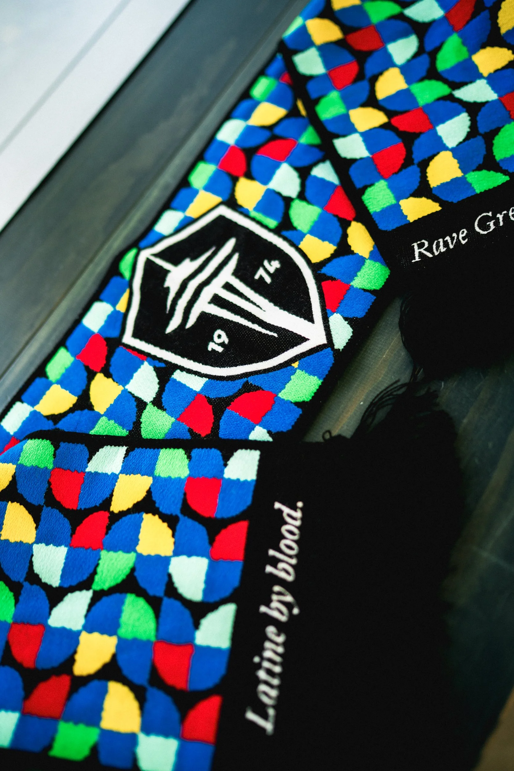 Colorful patterned fabric with a black patch that has a white shield-shaped logo with the numbers 19 and 74 inside and a stylized design. The fabric also displays the text "Lacing by blood.".