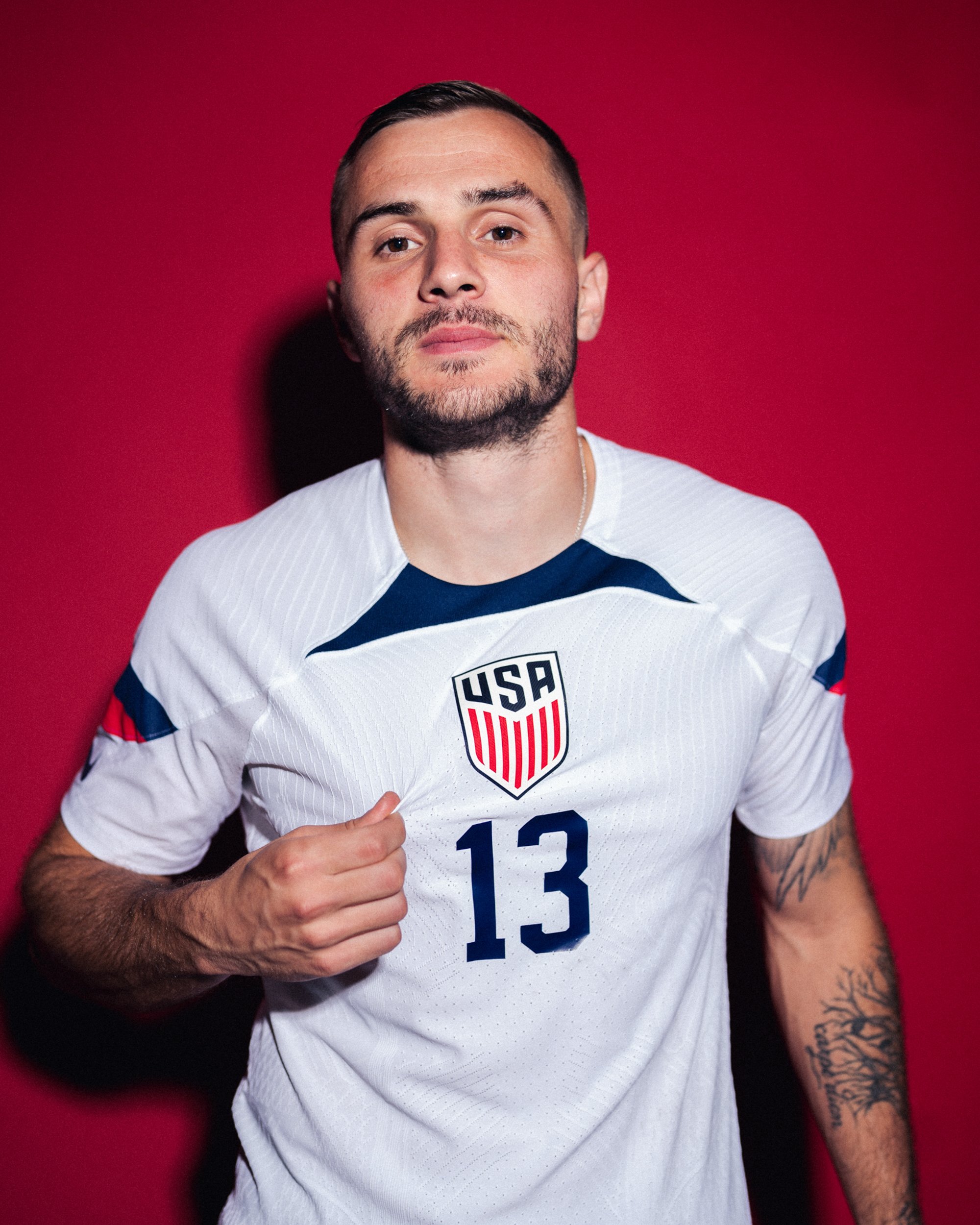 A man wearing a white sports jersey with USA emblem and the number 13, standing against a red background.