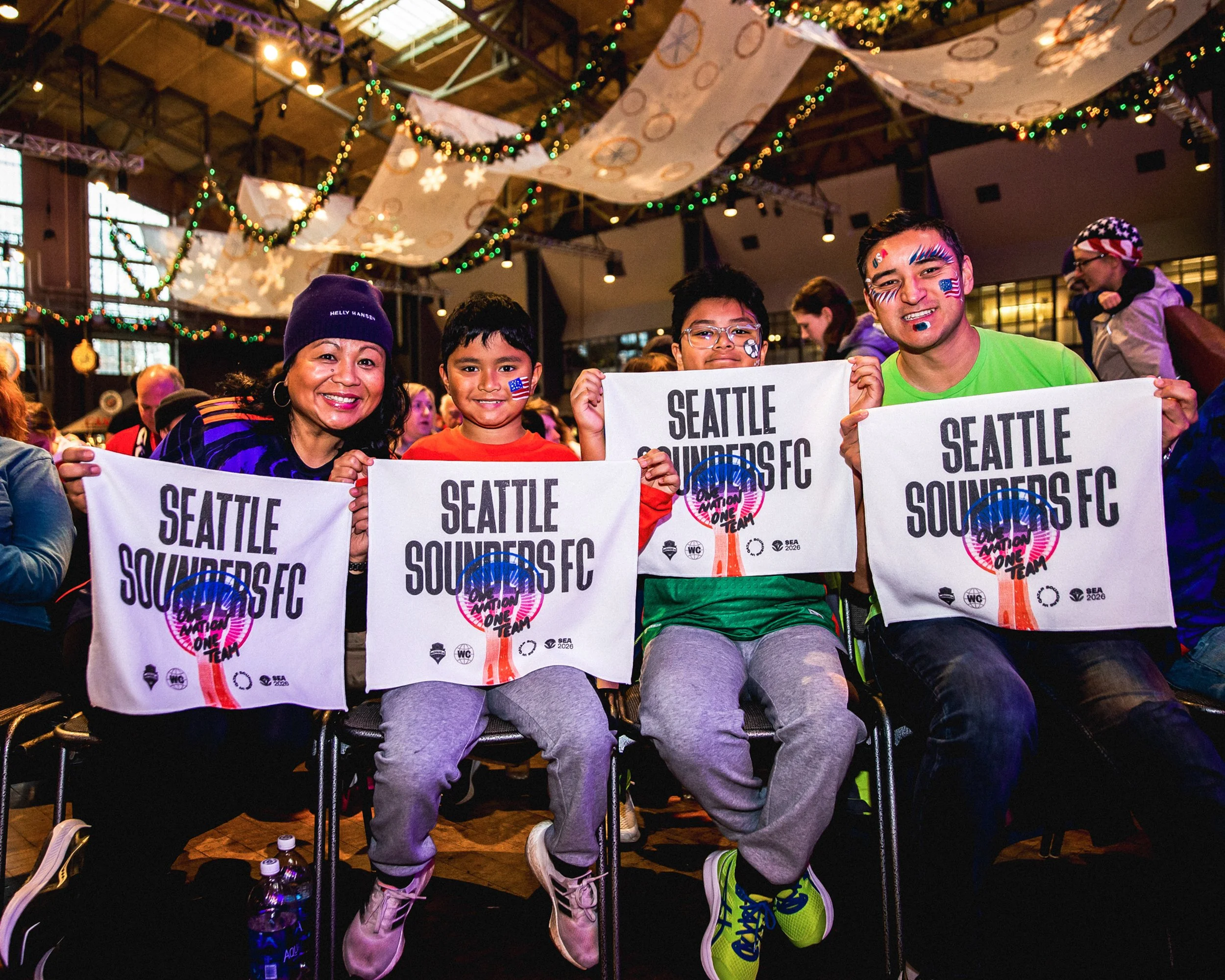 A group of four people, including two children and two adults, sitting in an indoor venue, holding flags with 'Seattle Sounders FC' and 'Major League Soccer' logos. They are smiling, with decorated face paint and some wearing American flags. The back