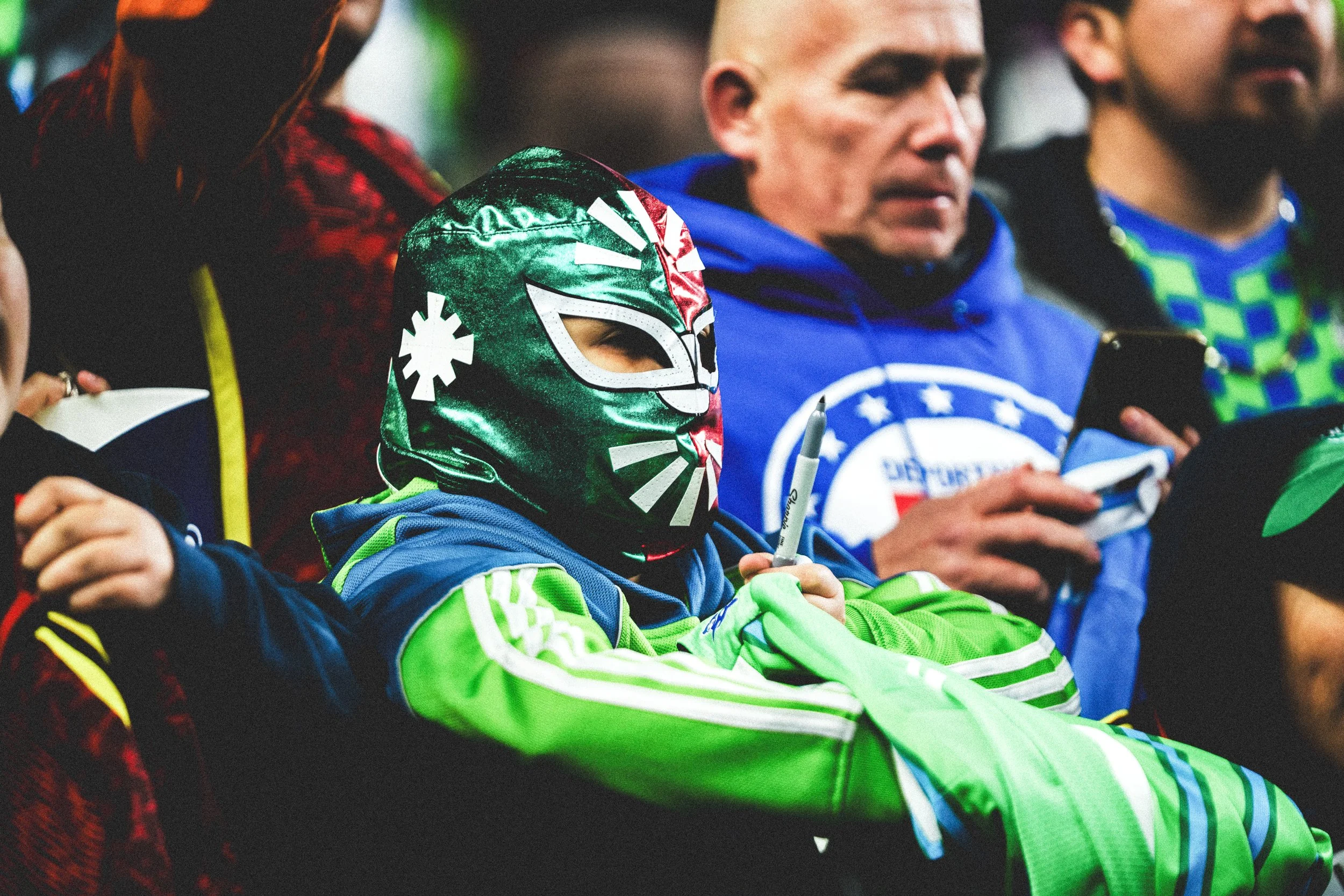 A group of people sitting in a row, some wearing colorful sports or team apparel. One person is wearing a shiny green mask resembling a luchador mask, with eye cutouts, and is holding a marker and a small object. Others are focused on their phones or