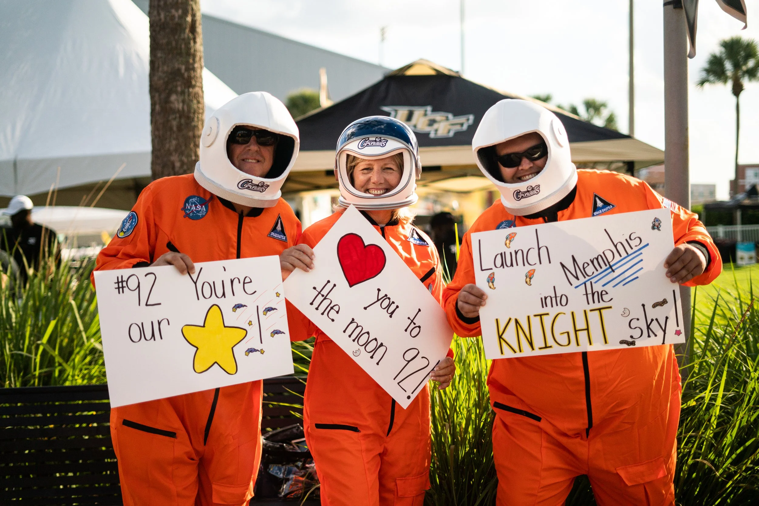 Three astronauts in orange space suits with helmets standing outdoors, holding signs with celebratory messages and decorative drawings.