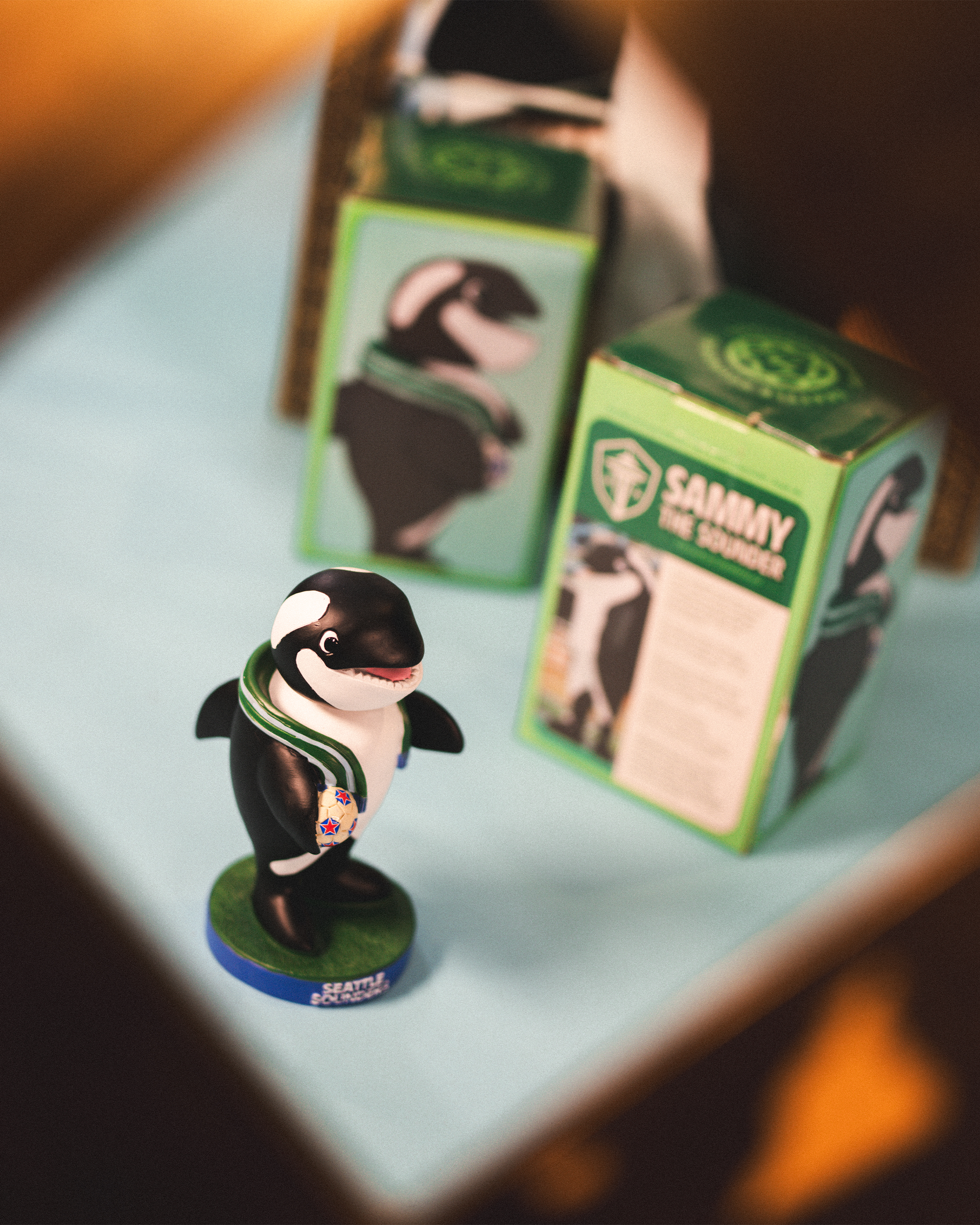 A small toy figure of a orca whale dressed as a race car driver, standing on a circular base with the text 'Seattle Seahawks.' Two boxes with the same orca image are in the background.