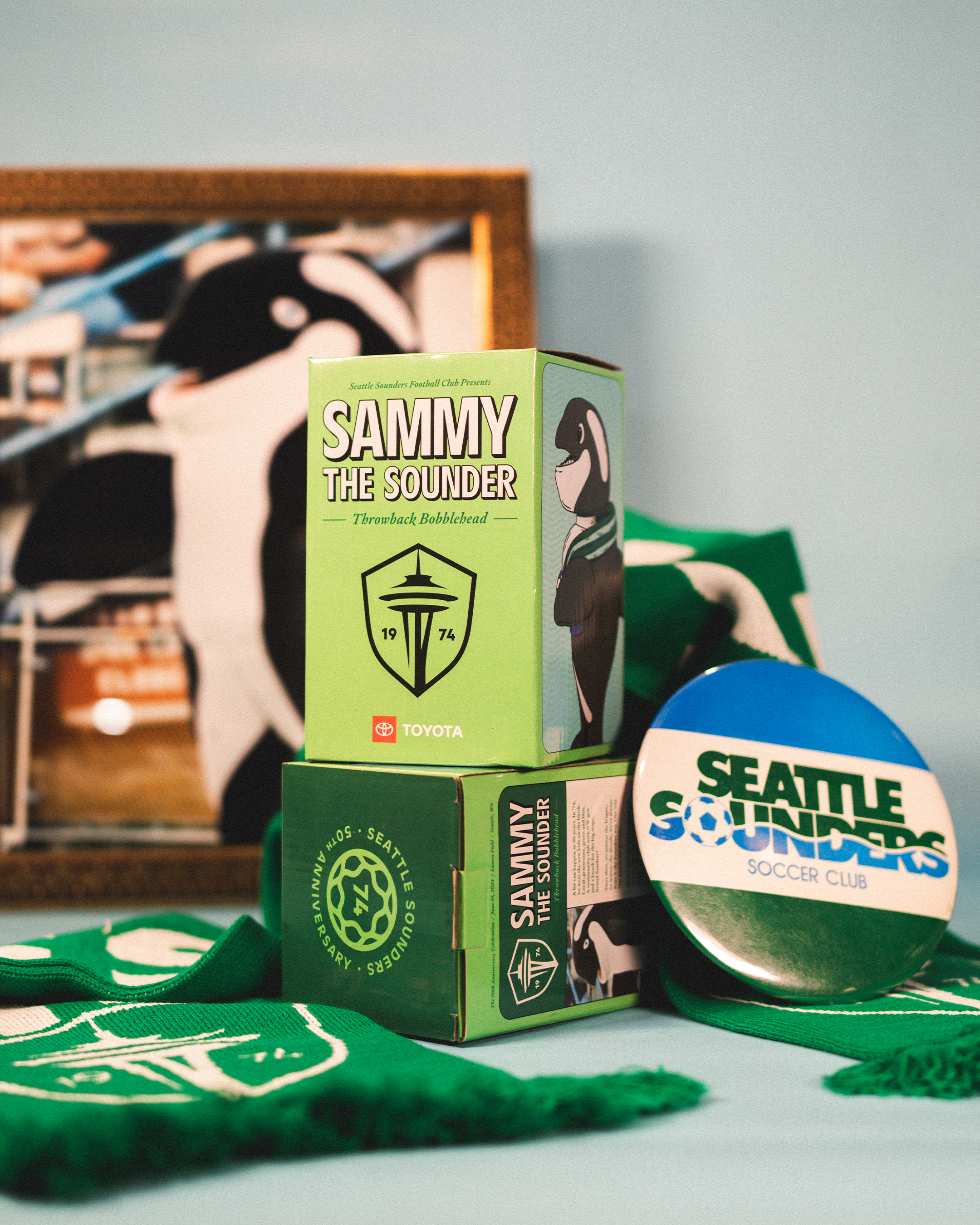 Seattle Sounders commemorative items including a green box, a round badge, and a green scarf, with a framed Sounders poster in the background.