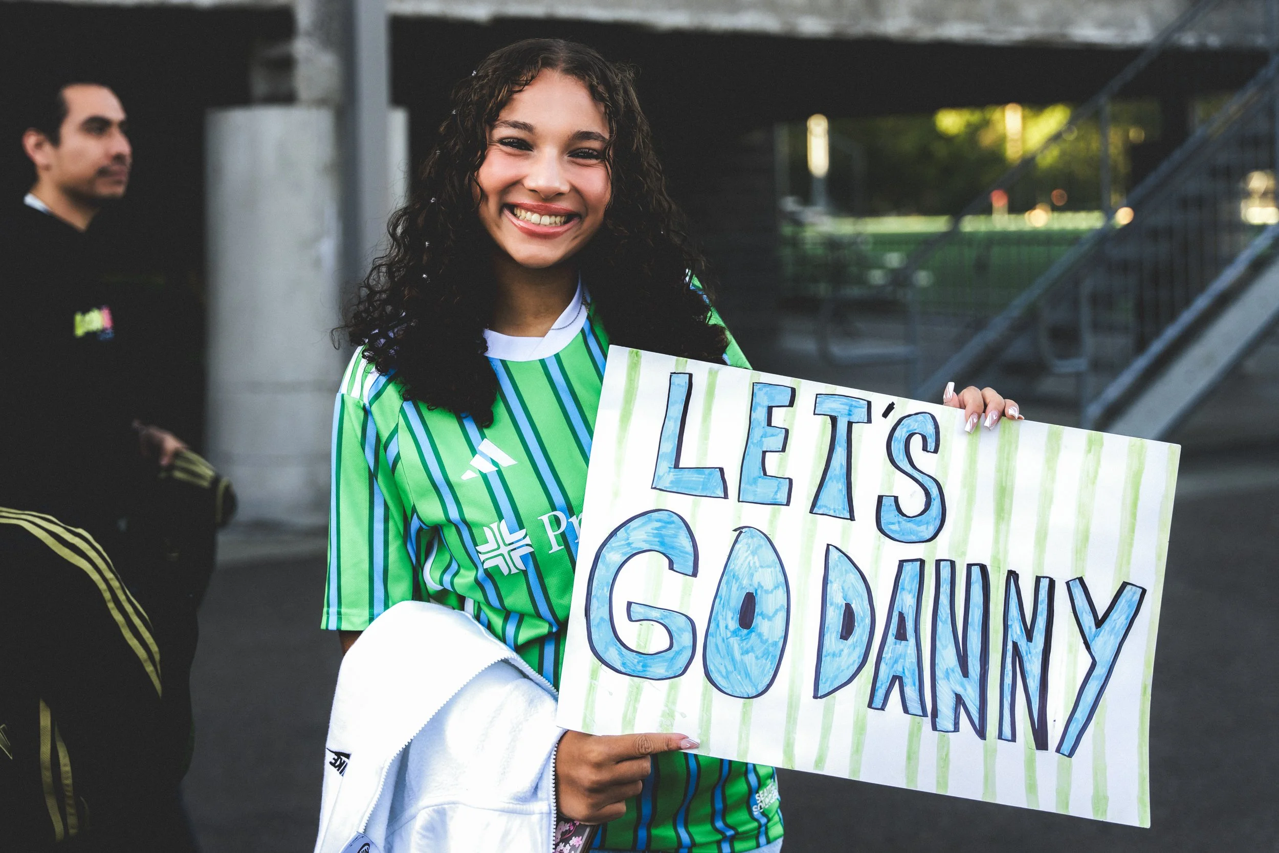 A smiling woman holding a sign that says 'Let's Go Danny' in a stadium or sports arena setting.