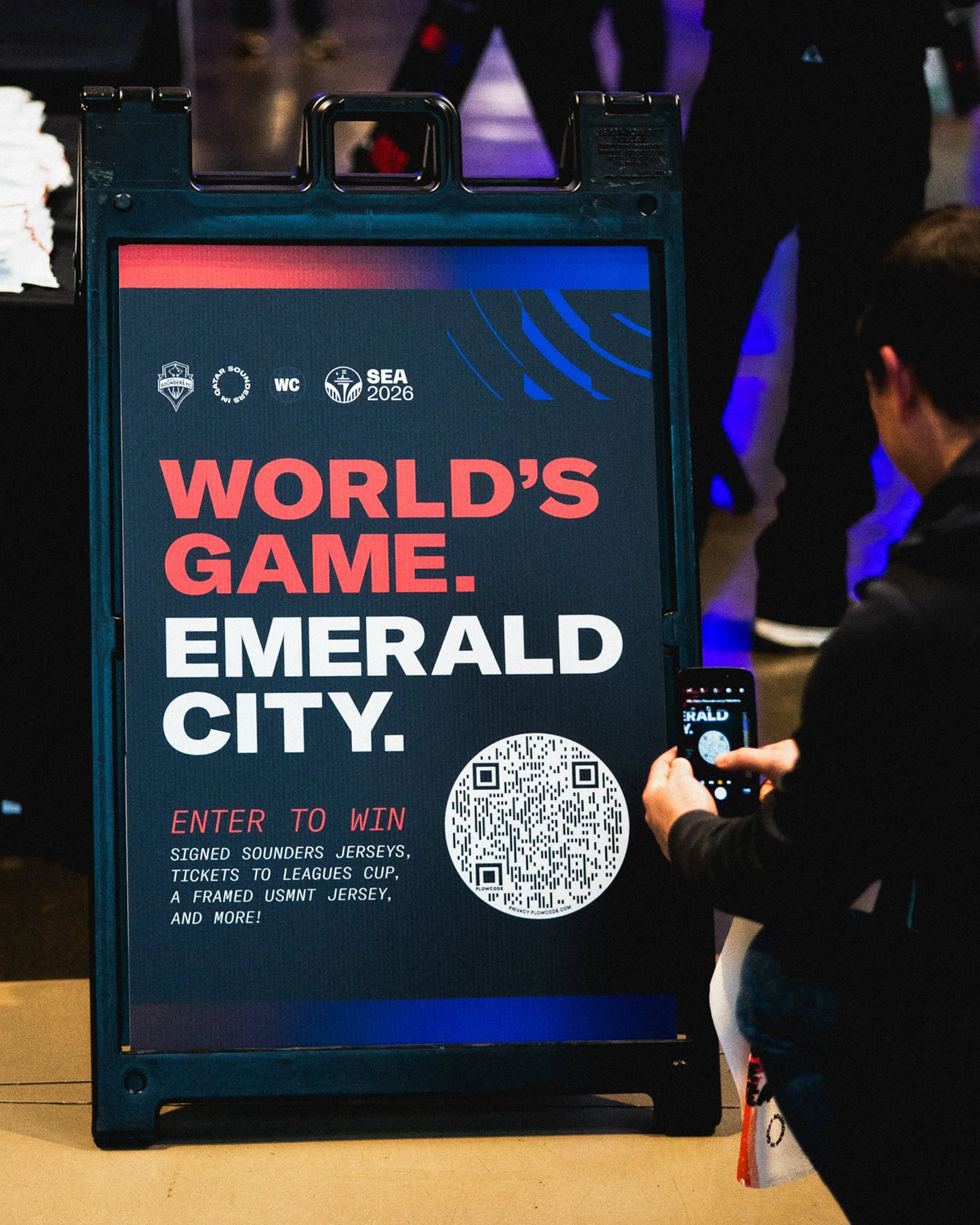 A person taking a photo of a sign promoting the 2026 World Cup in Emerald City with a smartphone. The sign has bold text and a QR code, mentioning a chance to win signed jerseys and tickets.