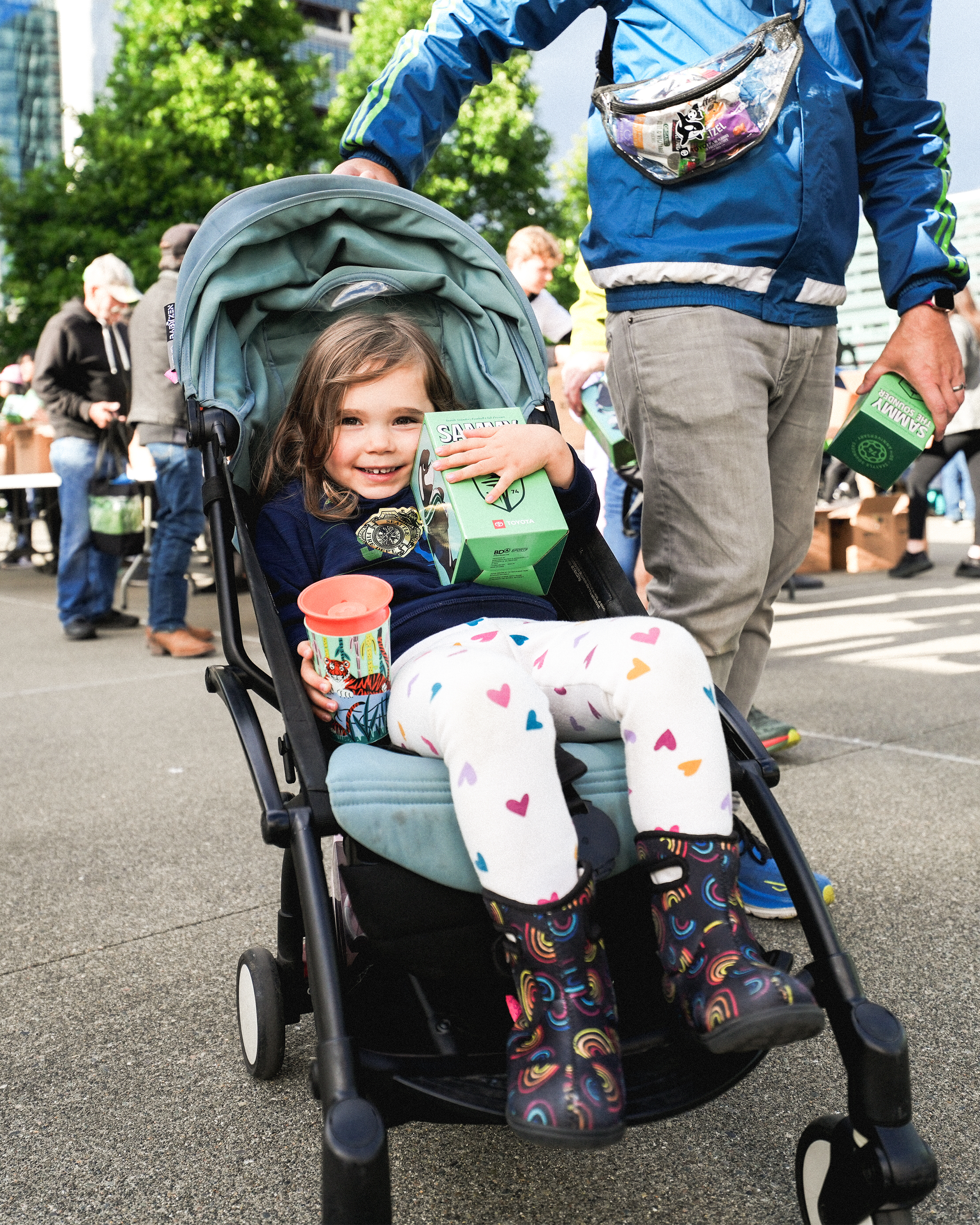 A young girl sitting in a stroller, holding a green toy car box and a colorful cup, smiling. She is at an outdoor event with people and tables in the background.