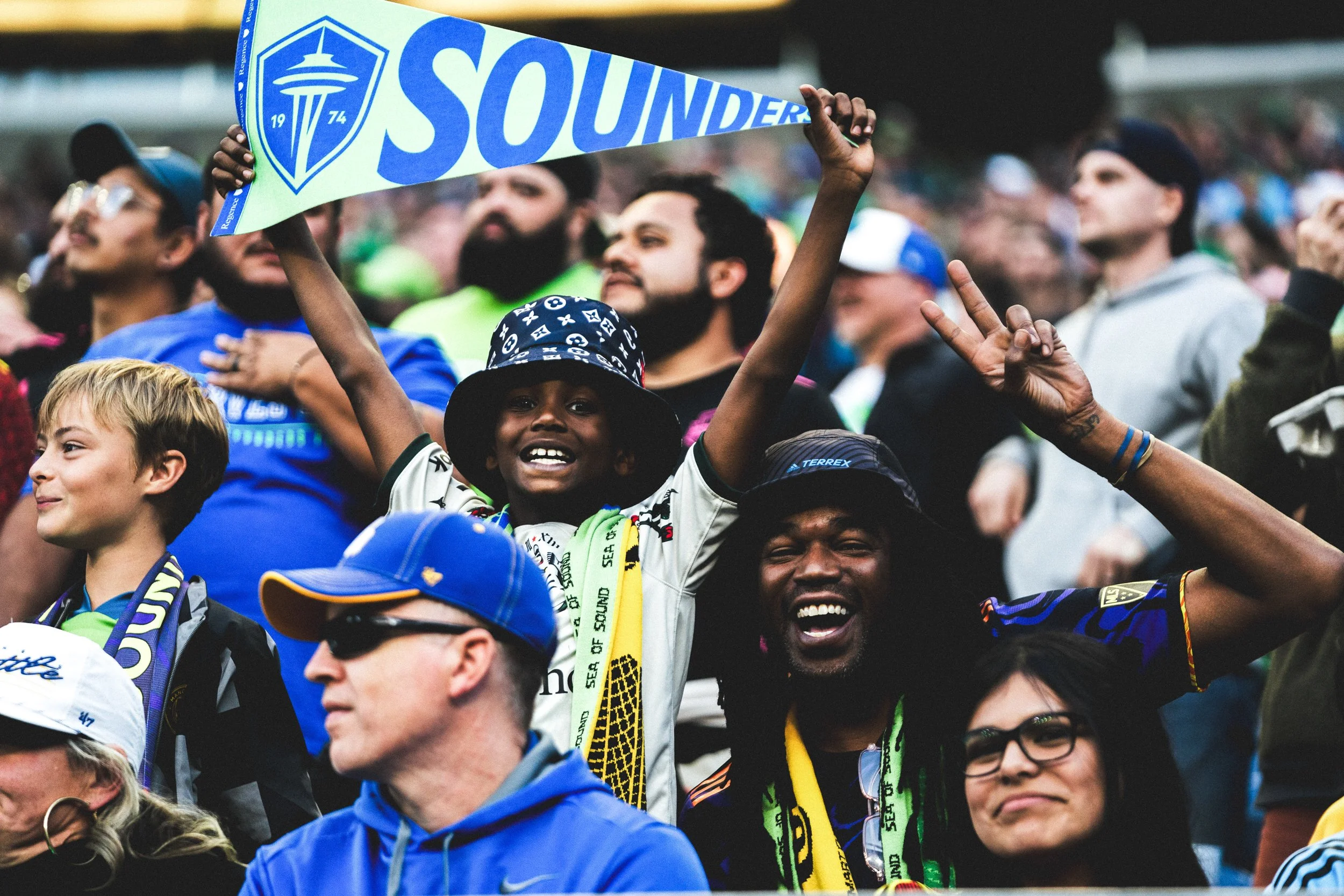 Fans at a sporting event cheering and waving team scarves, including a young boy holding a Seahawks scarf, with diverse faces and attire.