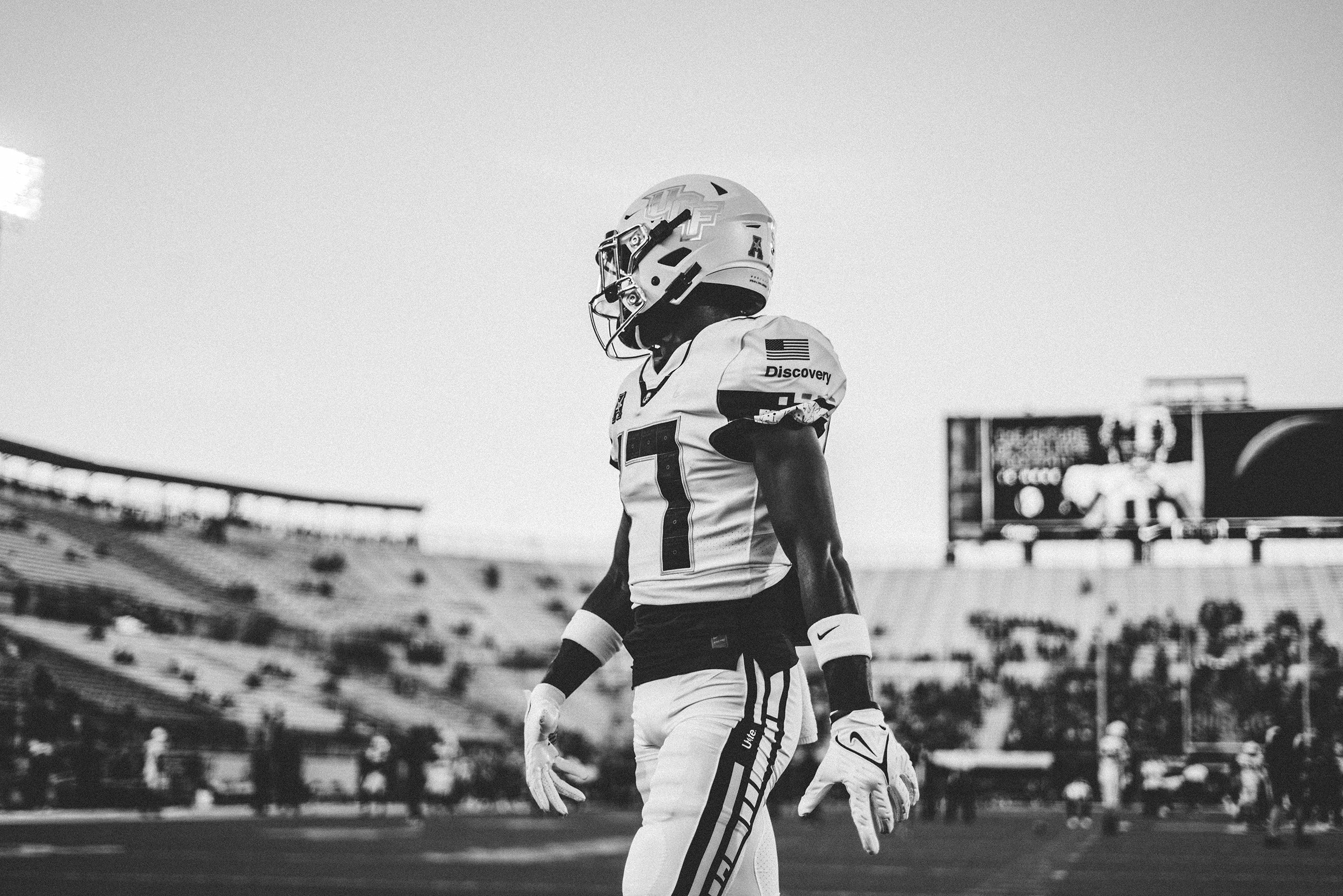 A football player in uniform standing on a football field in a stadium, wearing a helmet and gloves, with a large scoreboard in the background.
