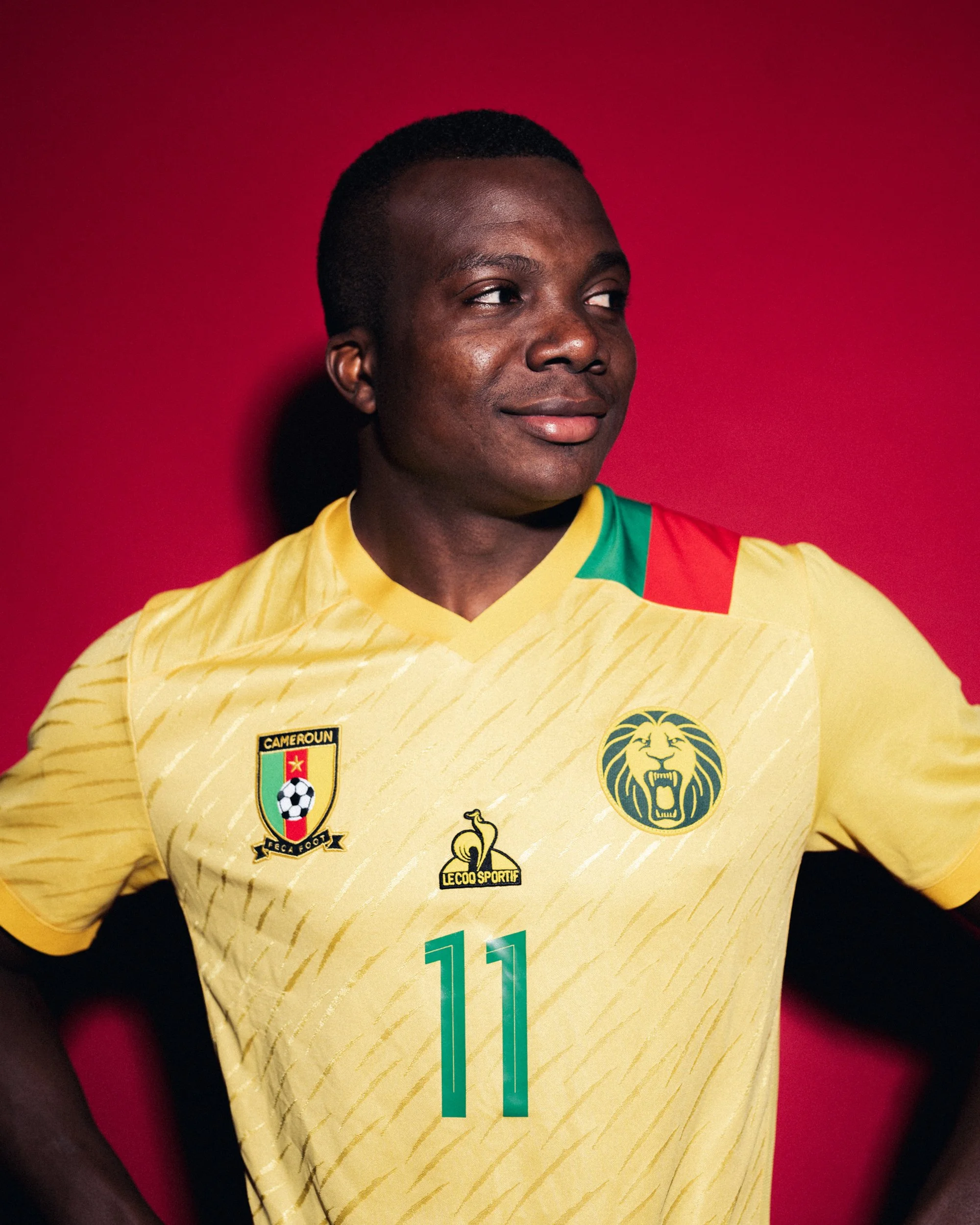 A male soccer player wearing a yellow Cameroon national team jersey with the number 11, standing against a red background.