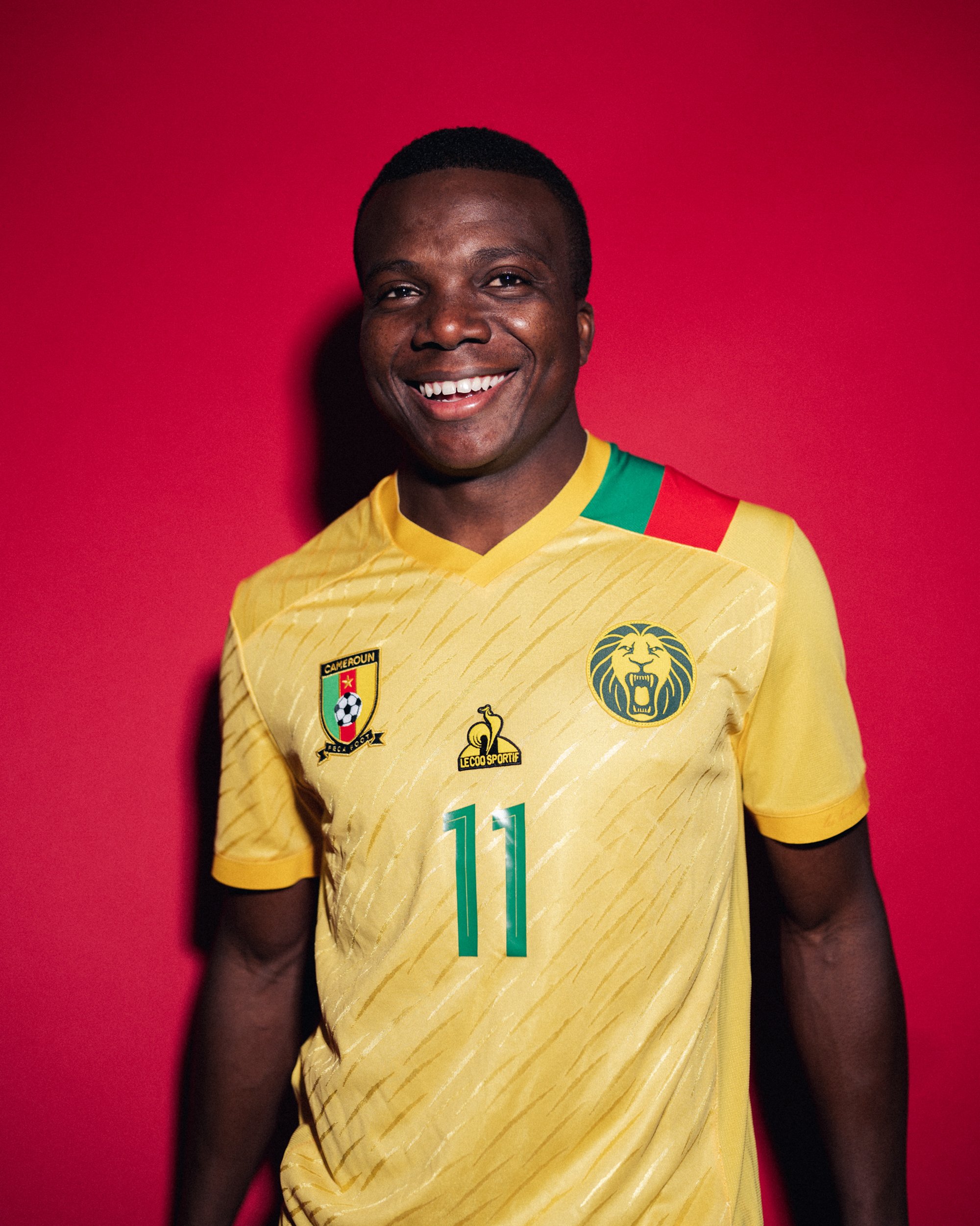 A smiling man wearing a Cameroon national football team jersey with the number 11, standing against a red background.
