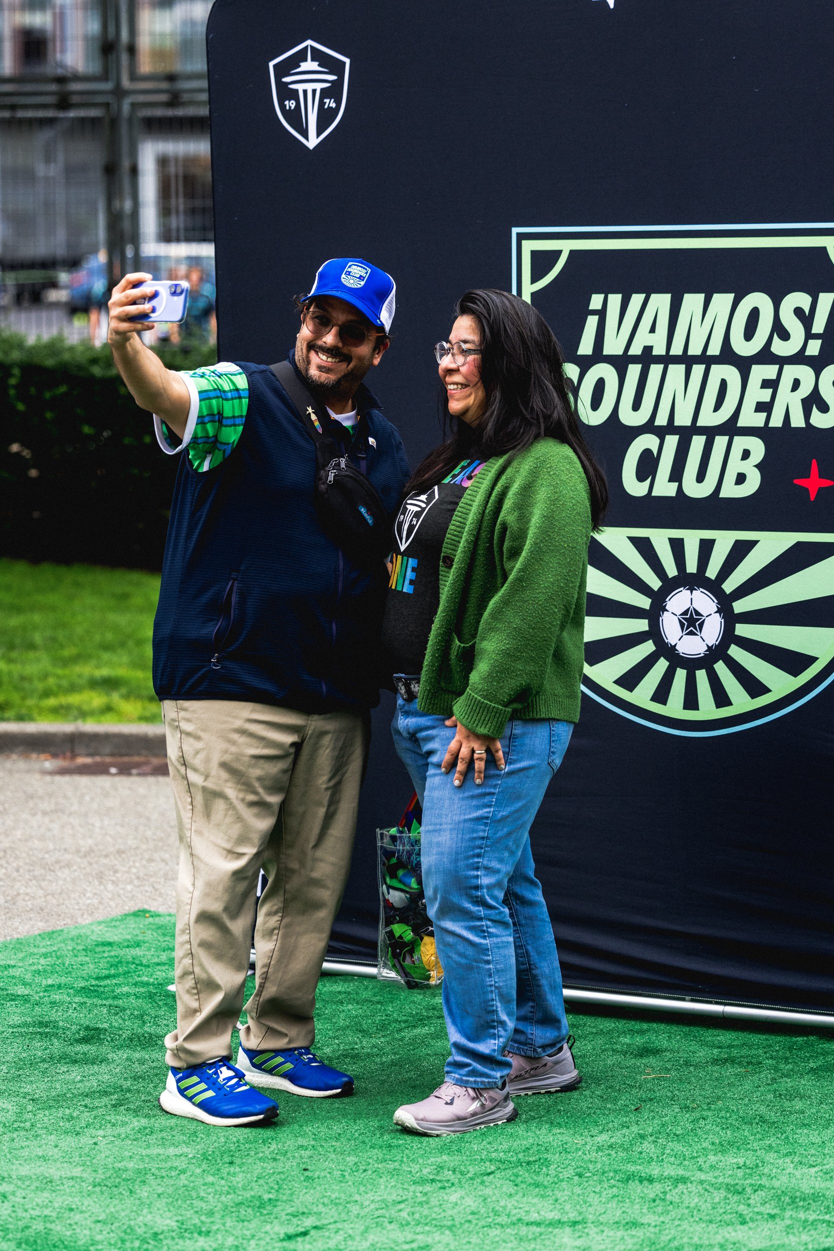 Two people taking a selfie in front of a sign that says "¡VAMOS! FOUNDERS CLUB" at a Seattle Sounders soccer event. The man is wearing a blue cap and glasses, and the woman has long dark hair and is wearing a green cardigan. They are both smiling.