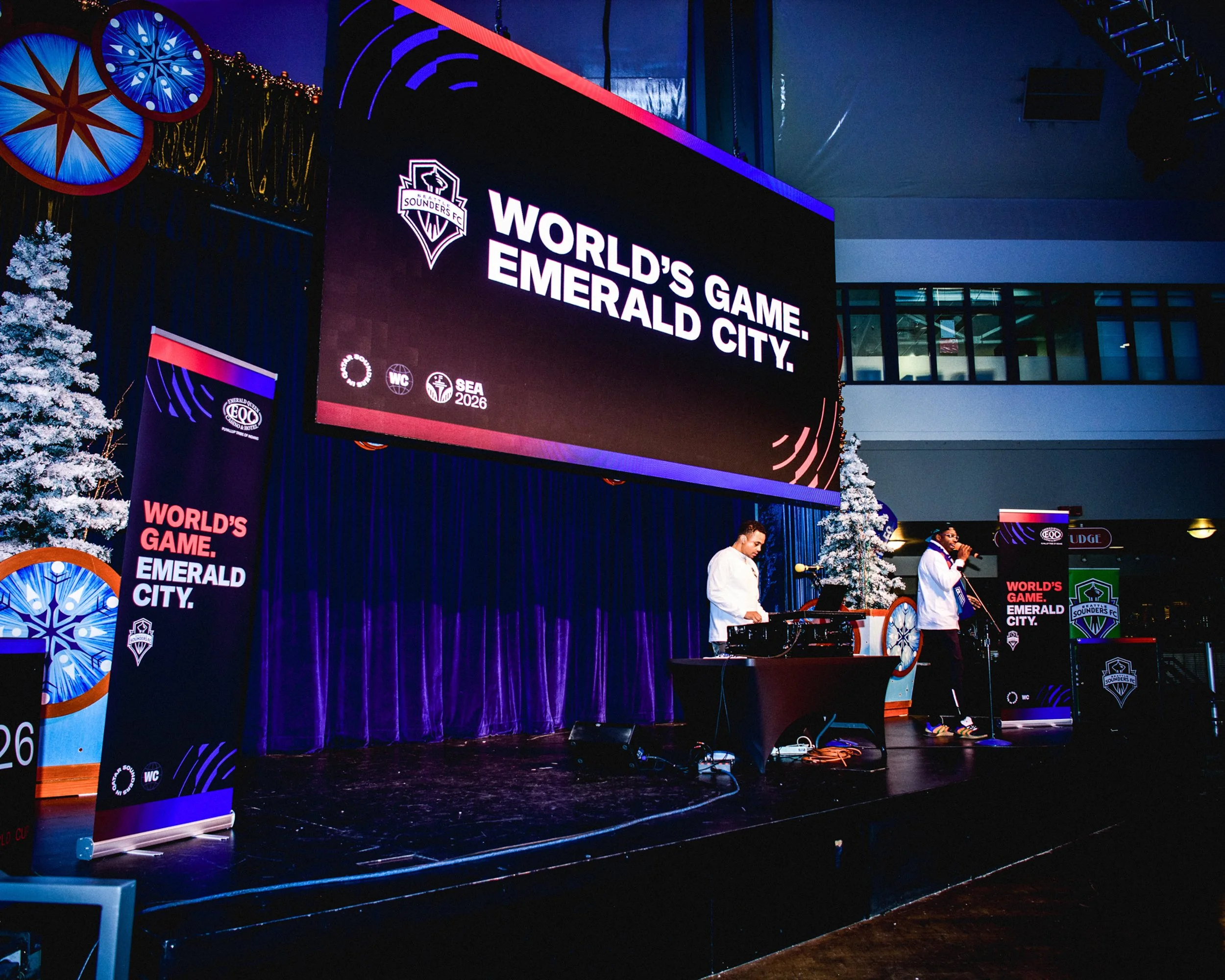Stage with large screen displaying logo and text 'World's Game. Emerald City.' at an event, with two performers or speakers on stage, decorated with Christmas trees and banners.