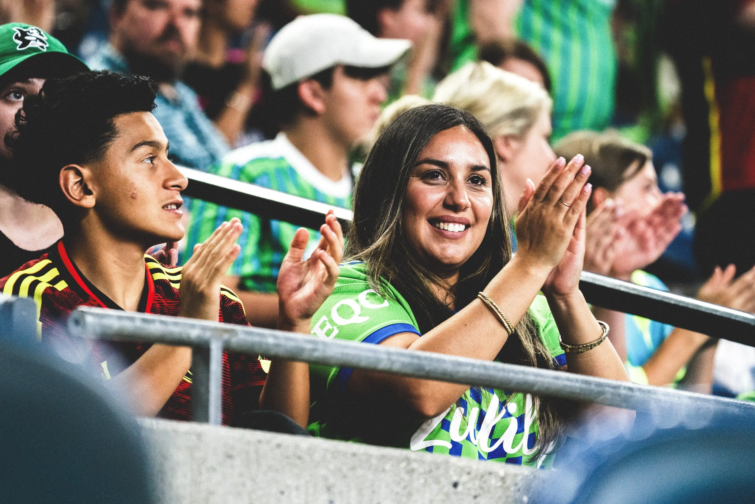 People sitting in stadium, clapping and cheering at a sporting event, woman smiling in foreground.