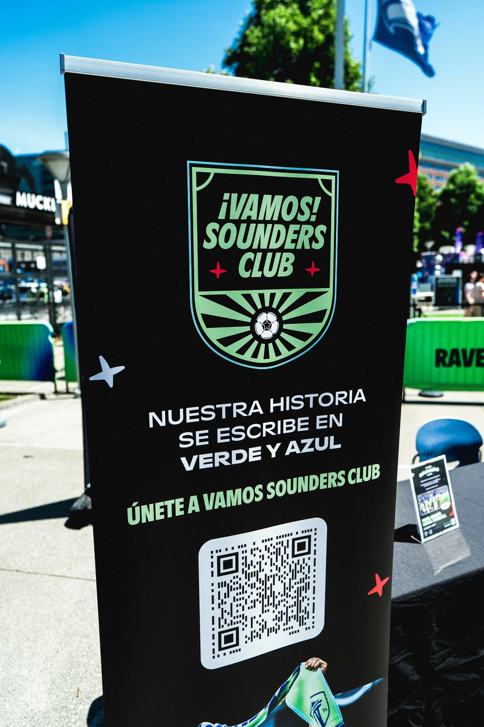 A black vertical banner for VAMOS! SOUNDER'S CLUB with the club's logo at the top, Spanish text underneath, a QR code below the text, and an image of a green and blue scarf at the bottom.