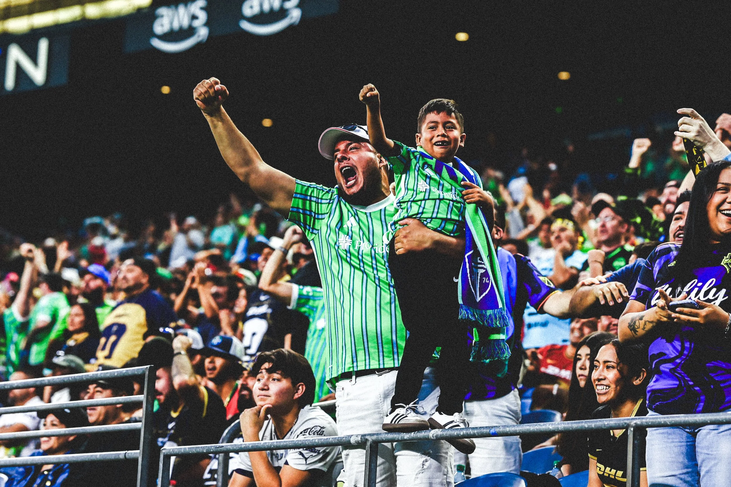 Fans cheering at a sports event, with a man holding a young boy on his shoulders, both wearing green and blue team jerseys, celebrating excitedly.
