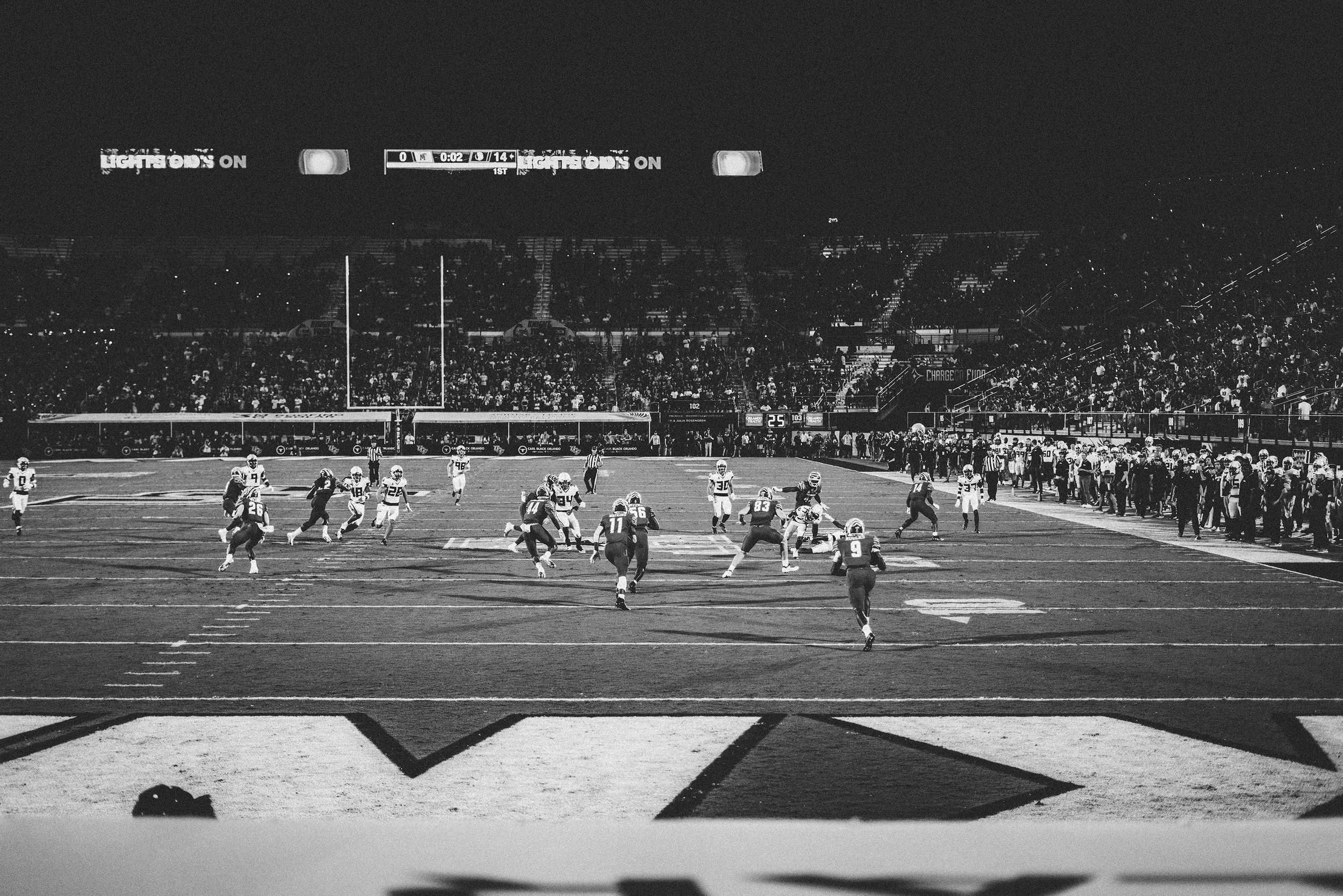 Black and white photo of an American football game at night, with players on the field, a crowd in the stands, and a scoreboard overhead.