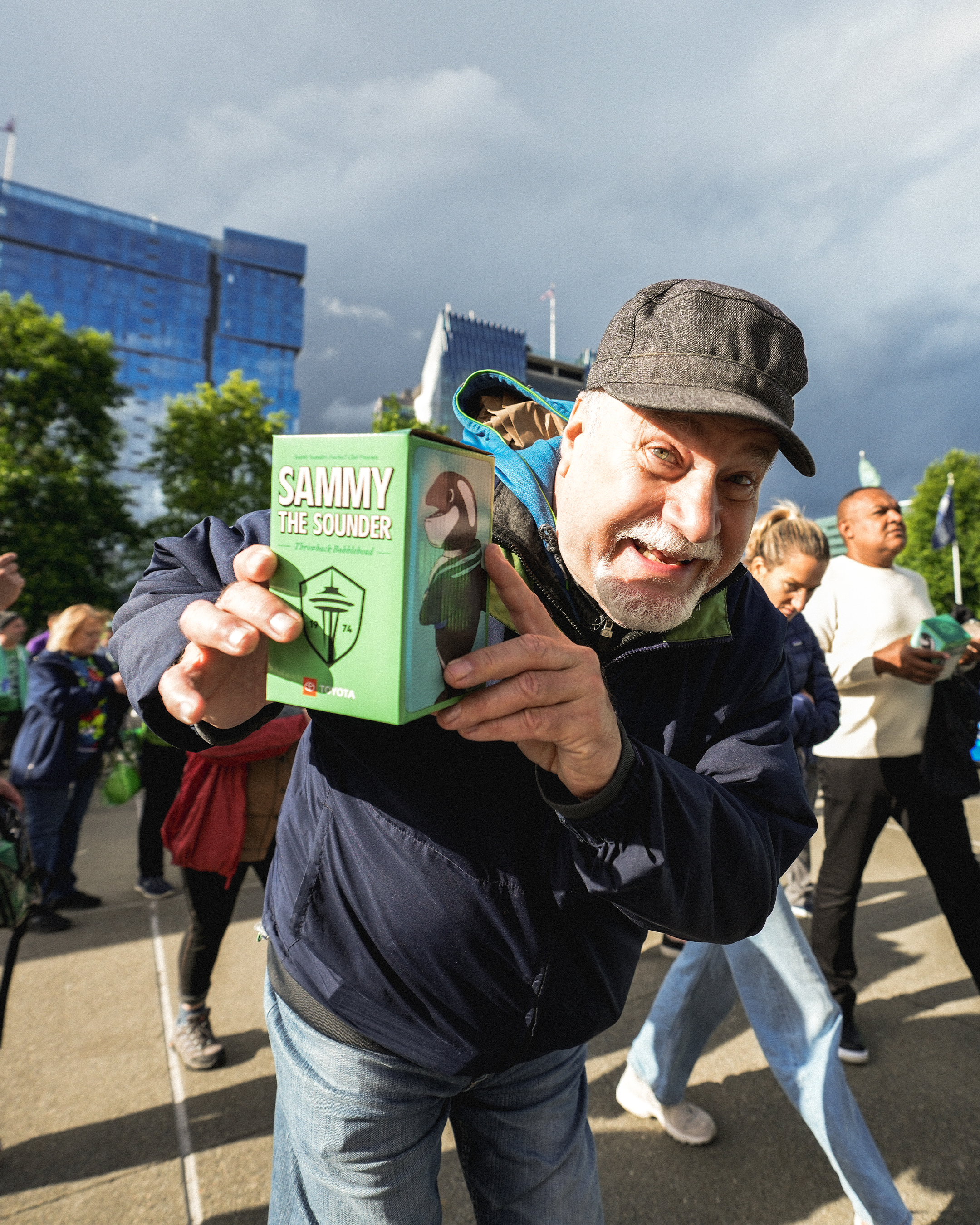A man holding a toy box labeled 'Sammy the Sounder' in a crowded outdoor setting with people and modern buildings in the background.