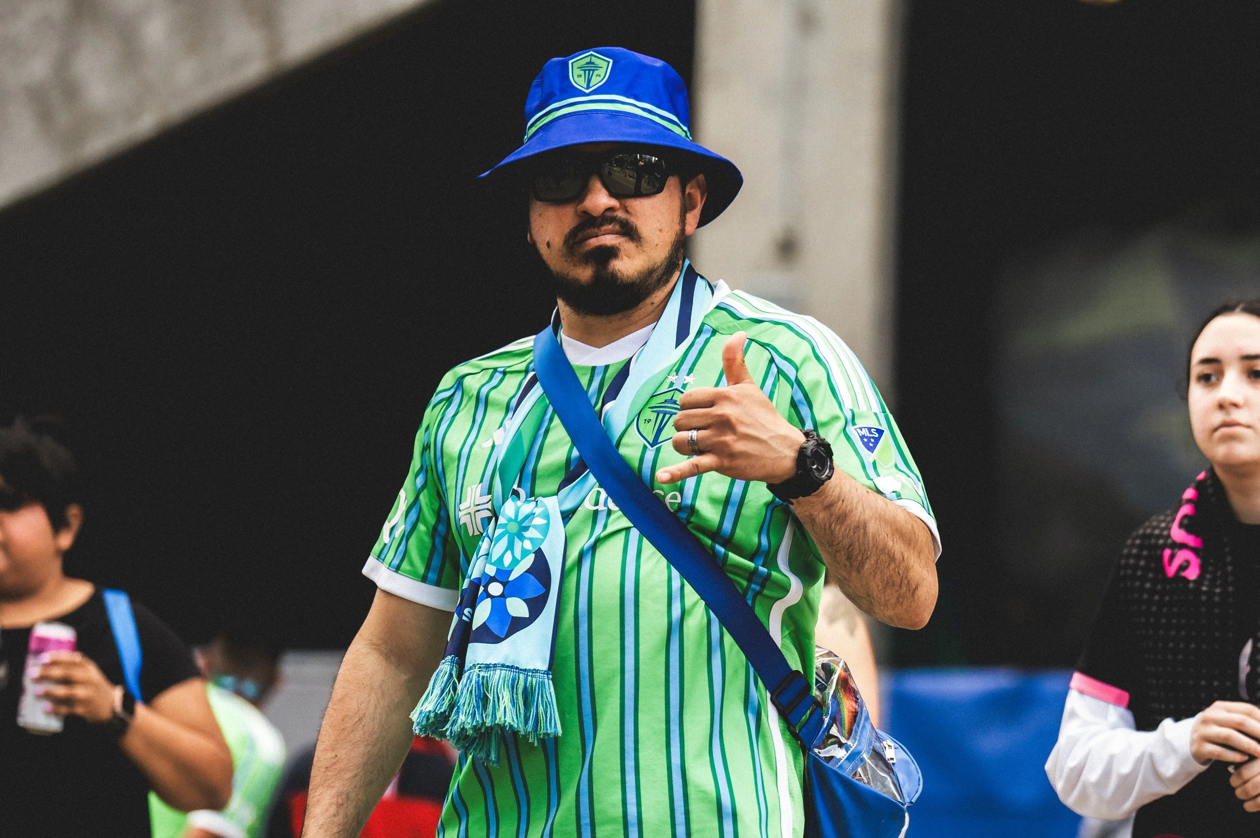 A man wearing a Seattle Sounders soccer jersey, sunglasses, hat, and a scarf around his neck showing a blue flower pattern, making a hand gesture, and carrying a blue shoulder bag, standing outdoors with other people in the background.