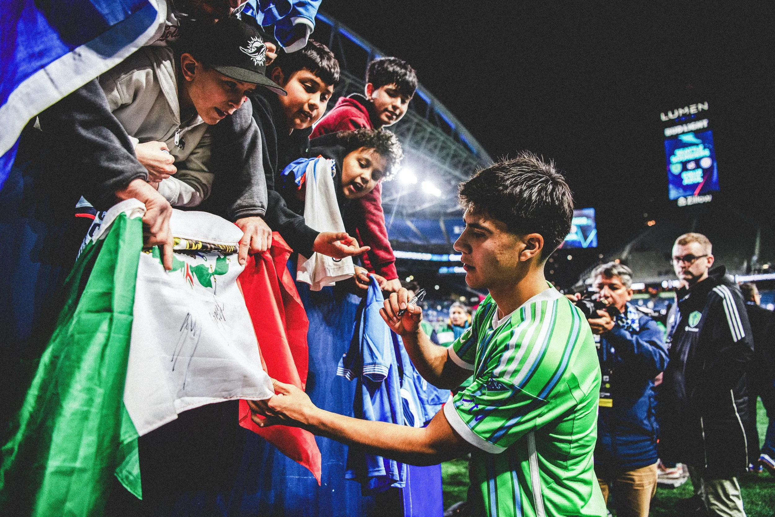 A young male soccer player in a green and white jersey signing autographs for children and fans behind a barrier at a stadium.