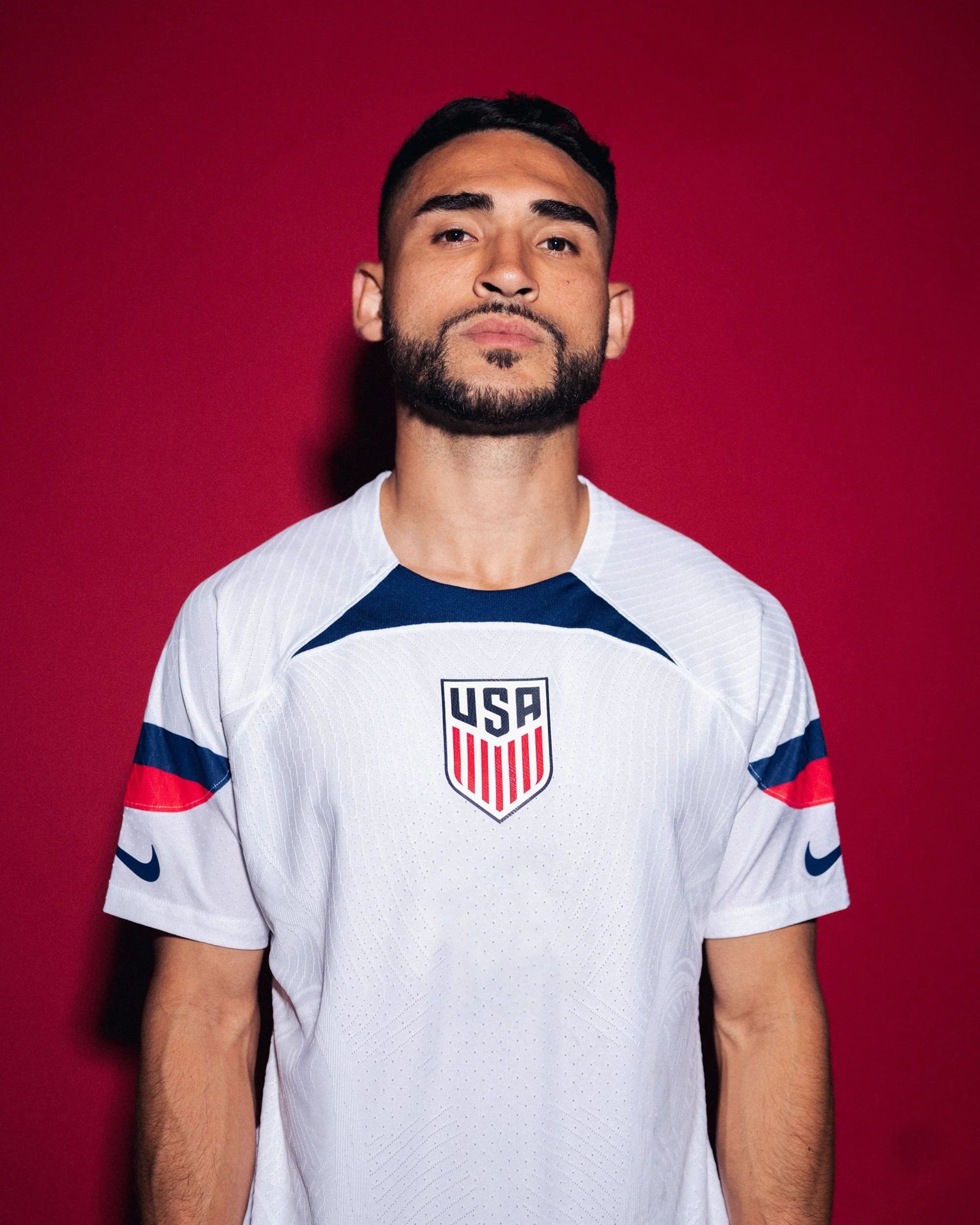 Man wearing a white USA soccer jersey with red, white, and blue accents, standing against a red background.