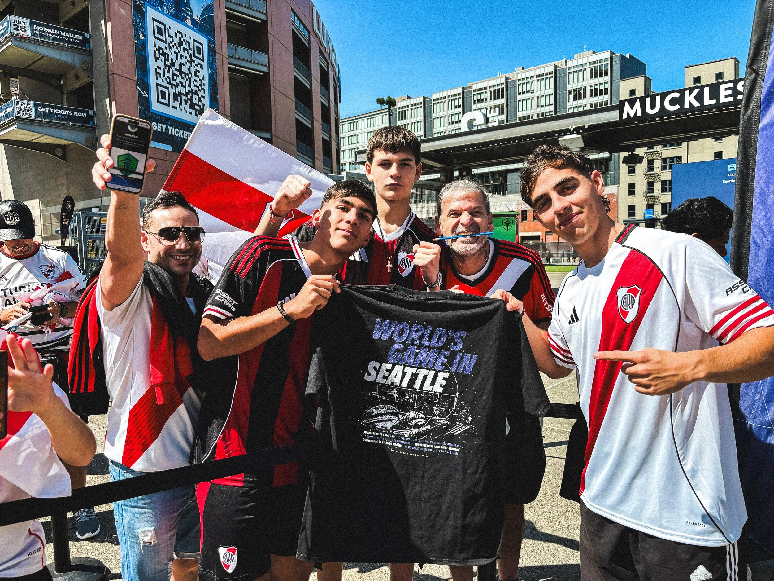 Group of football fans wearing River Plate jerseys, posing outdoors in Seattle with a flag and a black T-shirt that reads 'World's Game in Seattle', during daytime.