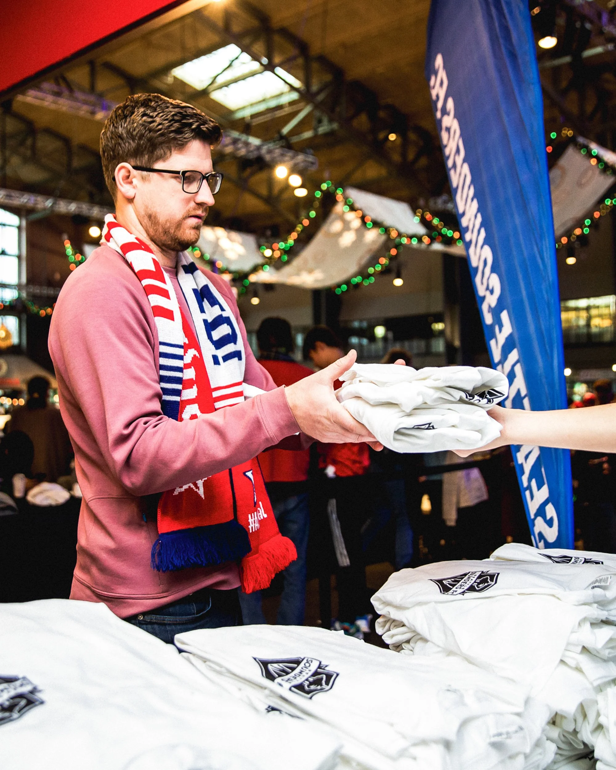 A man wearing glasses and a red sweatshirt exchanges a white t-shirt with a logo on it with someone off-camera. The scene appears to be at an indoor event with festive decorations, including string lights and a blue banner with white writing.
