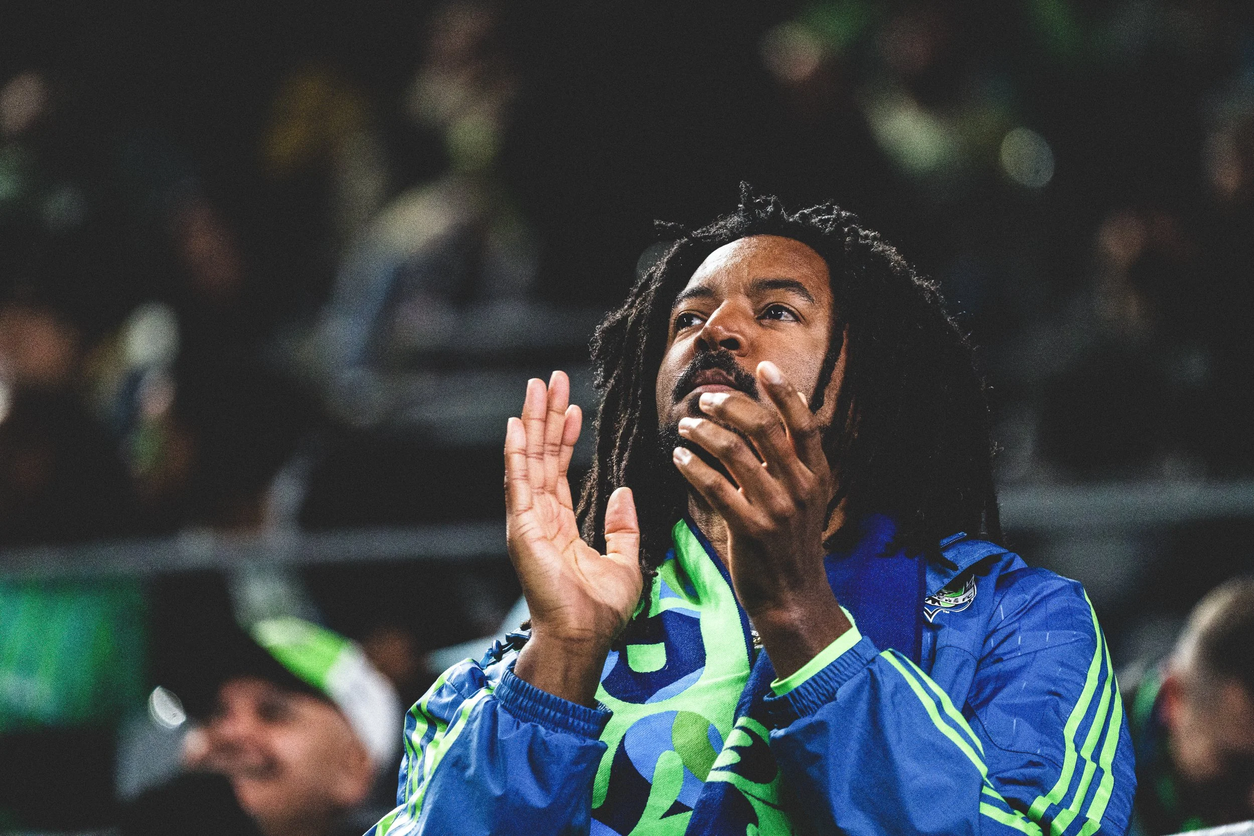 A man with dreadlocks wearing a Seattle Sounders FC soccer jersey at night, appearing to shout or cheer with his hands near his face, in a stadium or crowd setting.
