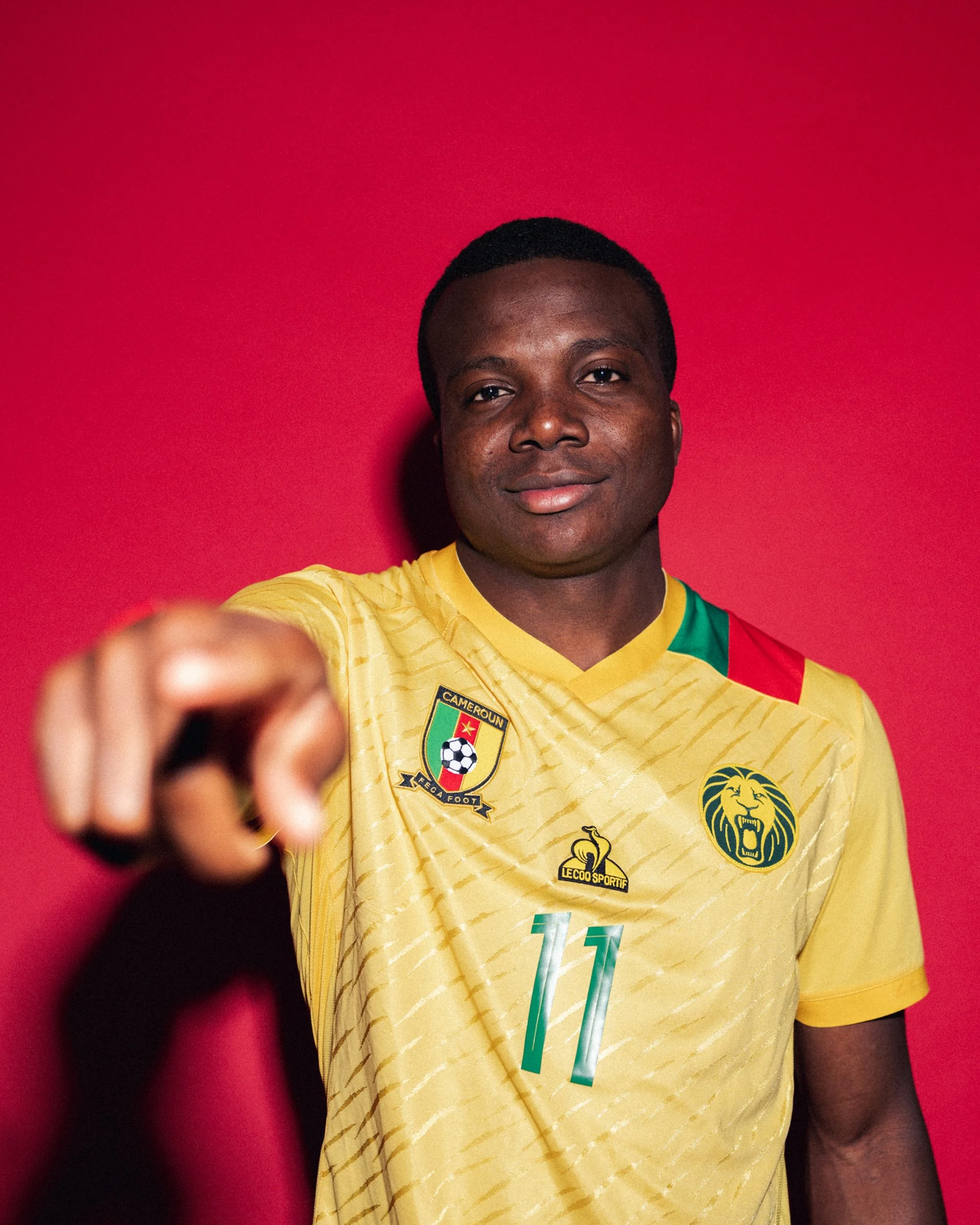 Portrait of a man in a yellow Cameroon national football team jersey pointing towards the camera, standing against a red background.